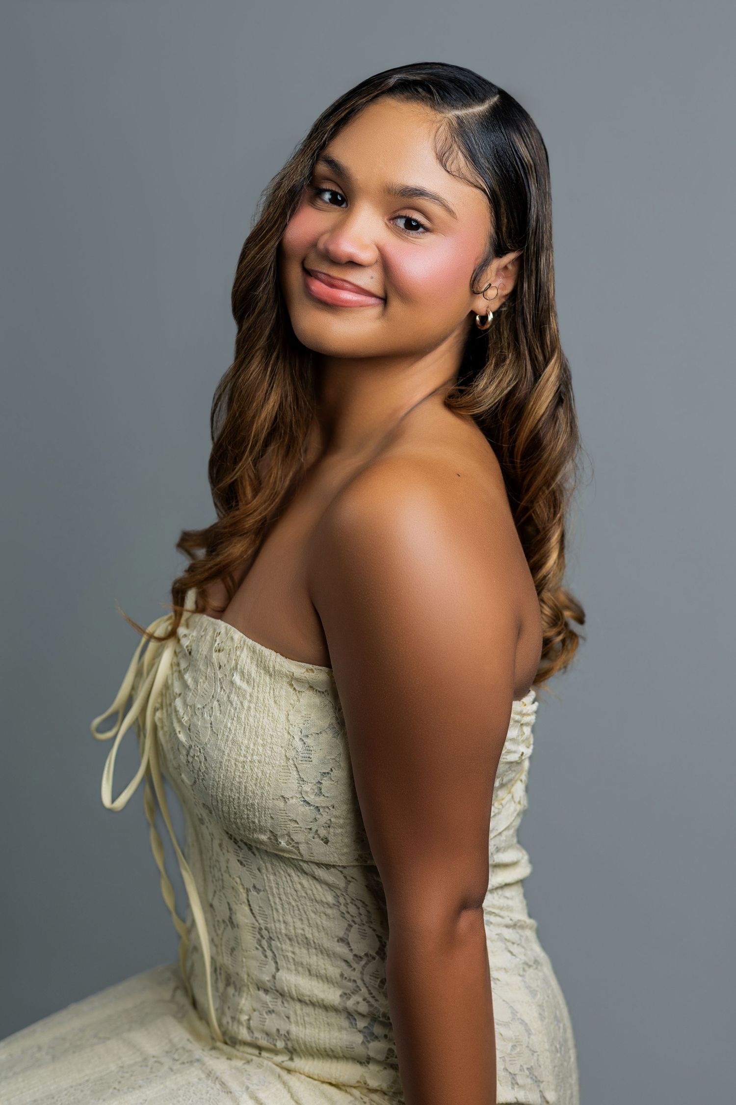 Young woman in strapless cream lace dress smiling over shoulder, studio portrait with gray background.