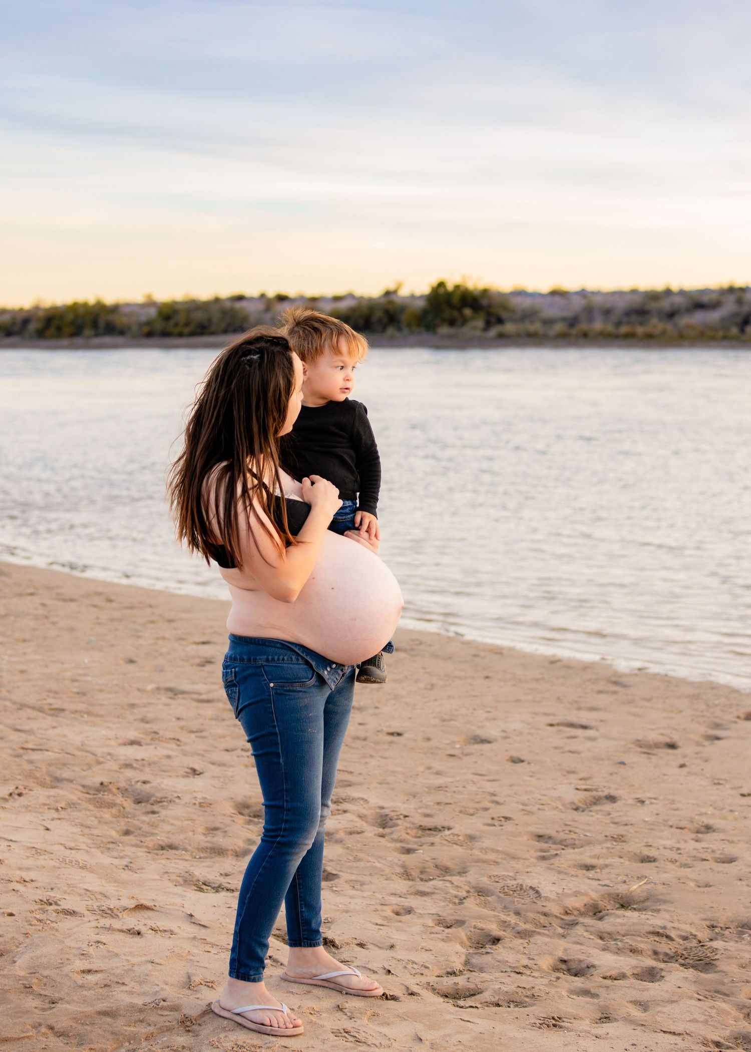 Pregnant mother holding toddler son on sandy beach at golden hour sunset.