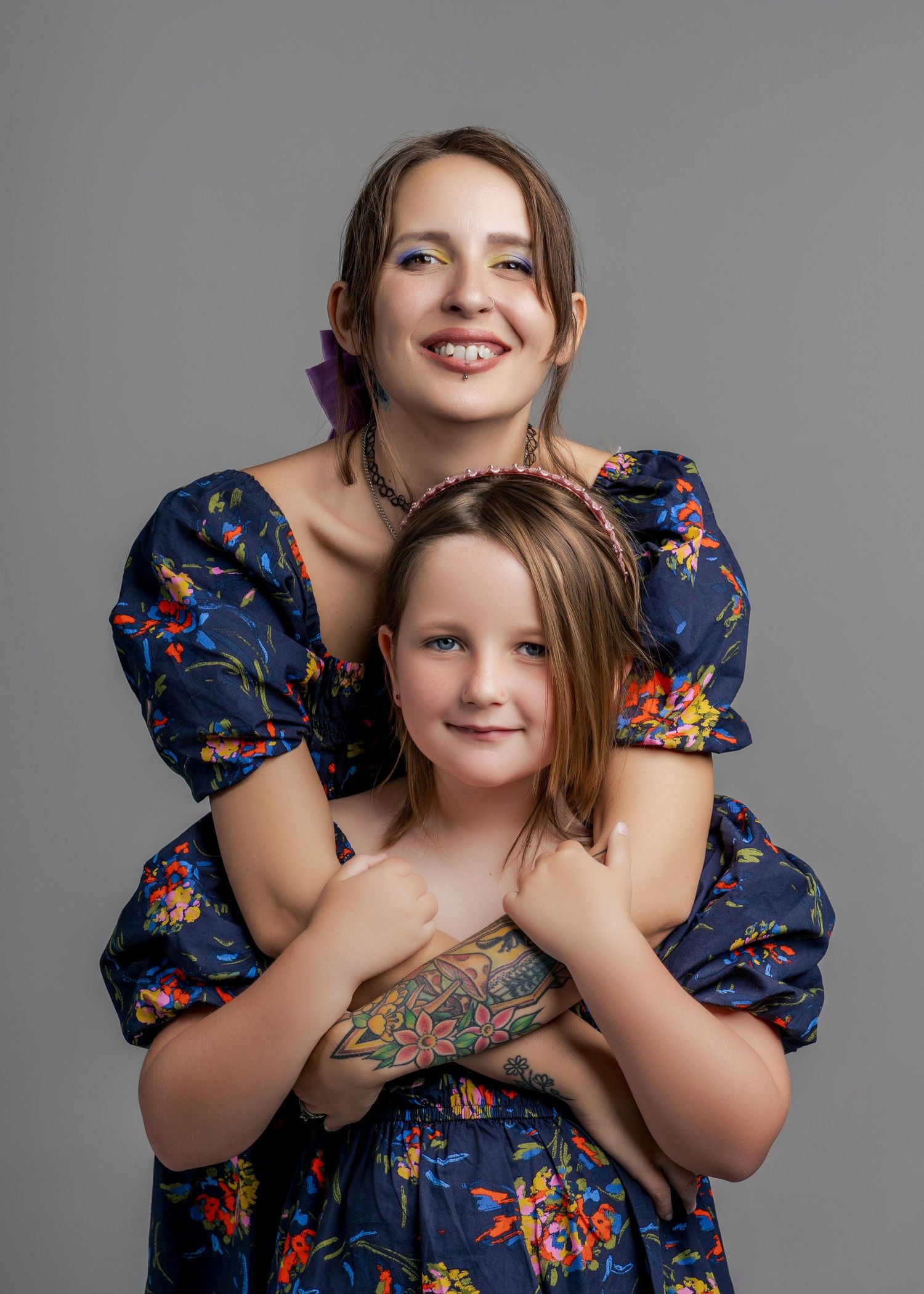 A smiling woman in a blue floral dress embraces a young girl in a warm studio portrait.