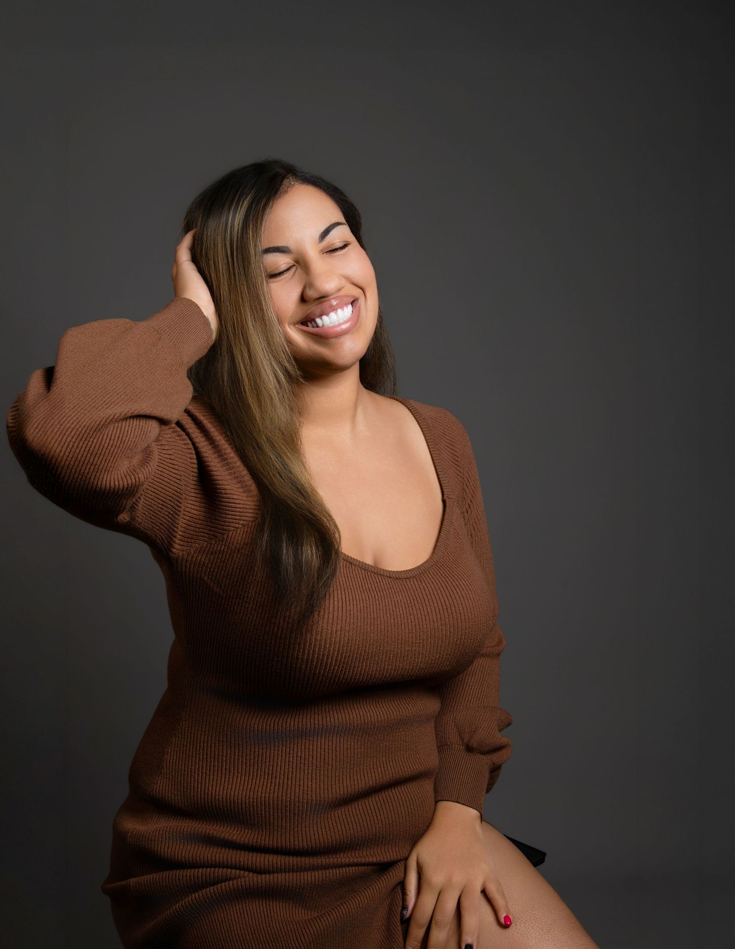 Joyful woman in brown sweater laughs with eyes closed while touching hair on gray background.