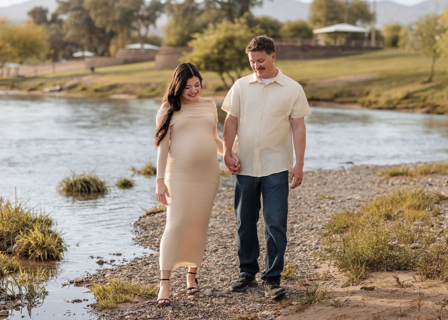 Pregnant couple holding hands walking along a scenic riverside path, woman in beige maternity dress.