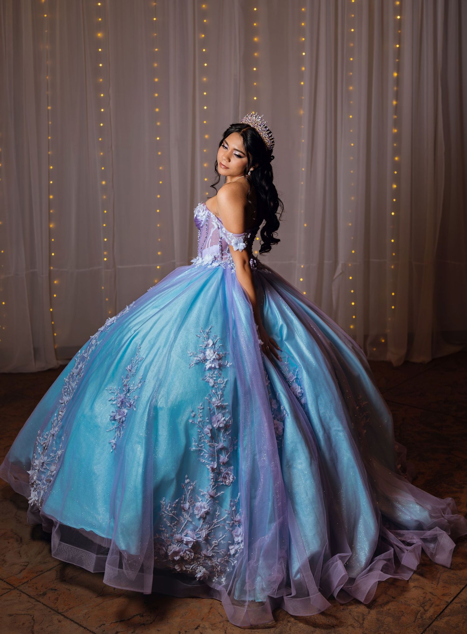 Elegant quinceañera in blue and purple ball gown with floral appliques and sparkling tiara posing.