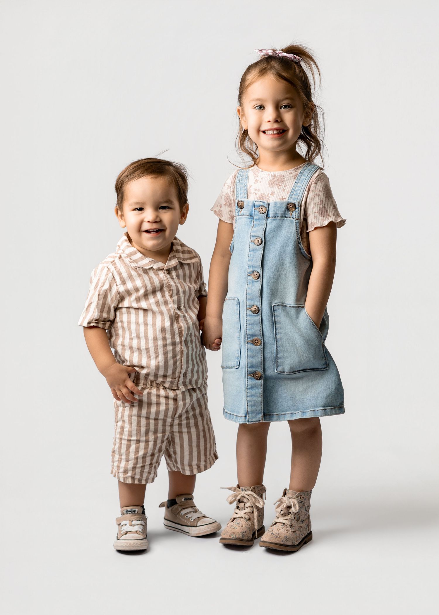Two smiling toddlers posing together, boy in striped shorts set and girl in denim overall dress with sneakers.
