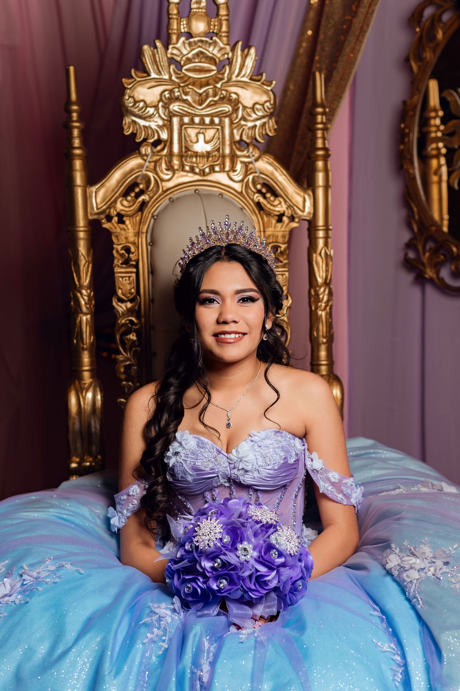 Smiling quinceañera in blue and purple gown holding purple bouquet seated on ornate golden throne.