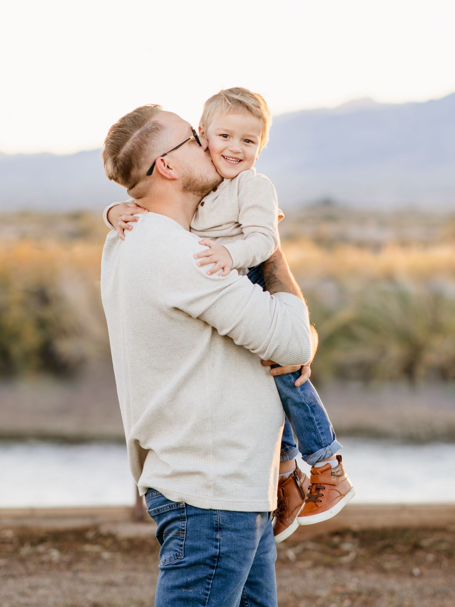 Father kisses smiling young son while holding him outdoors near a lake at golden hour.