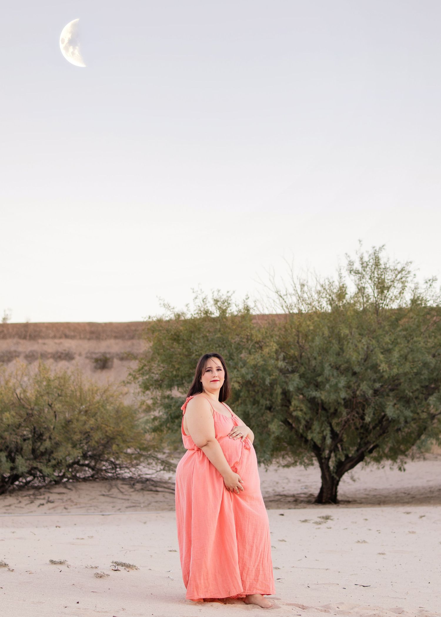 Pregnant woman in flowing coral dress stands in desert landscape under crescent moon at dusk.