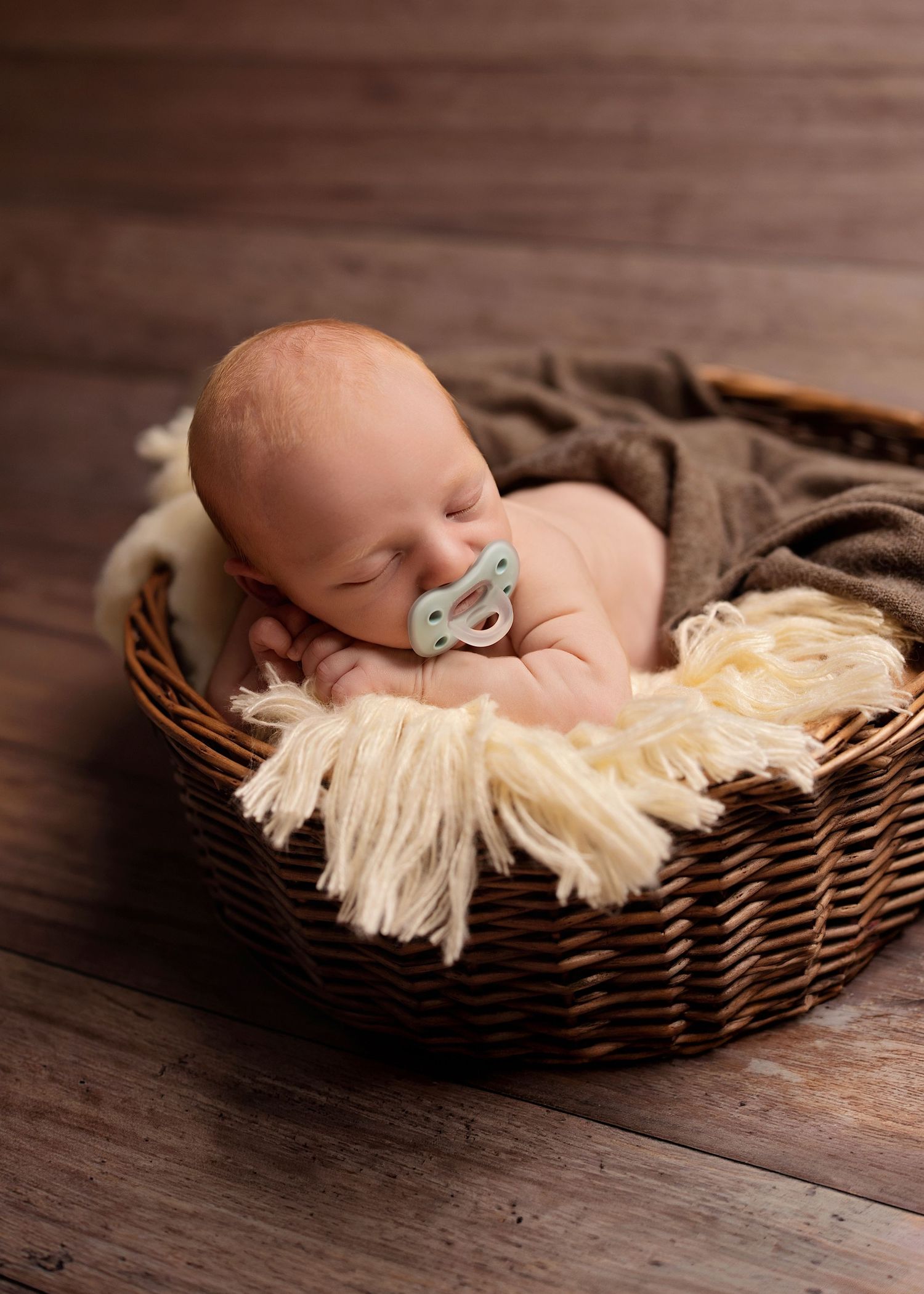 Sleeping newborn baby with pacifier rests peacefully in wicker basket with cream fringe blanket on wood floor.
