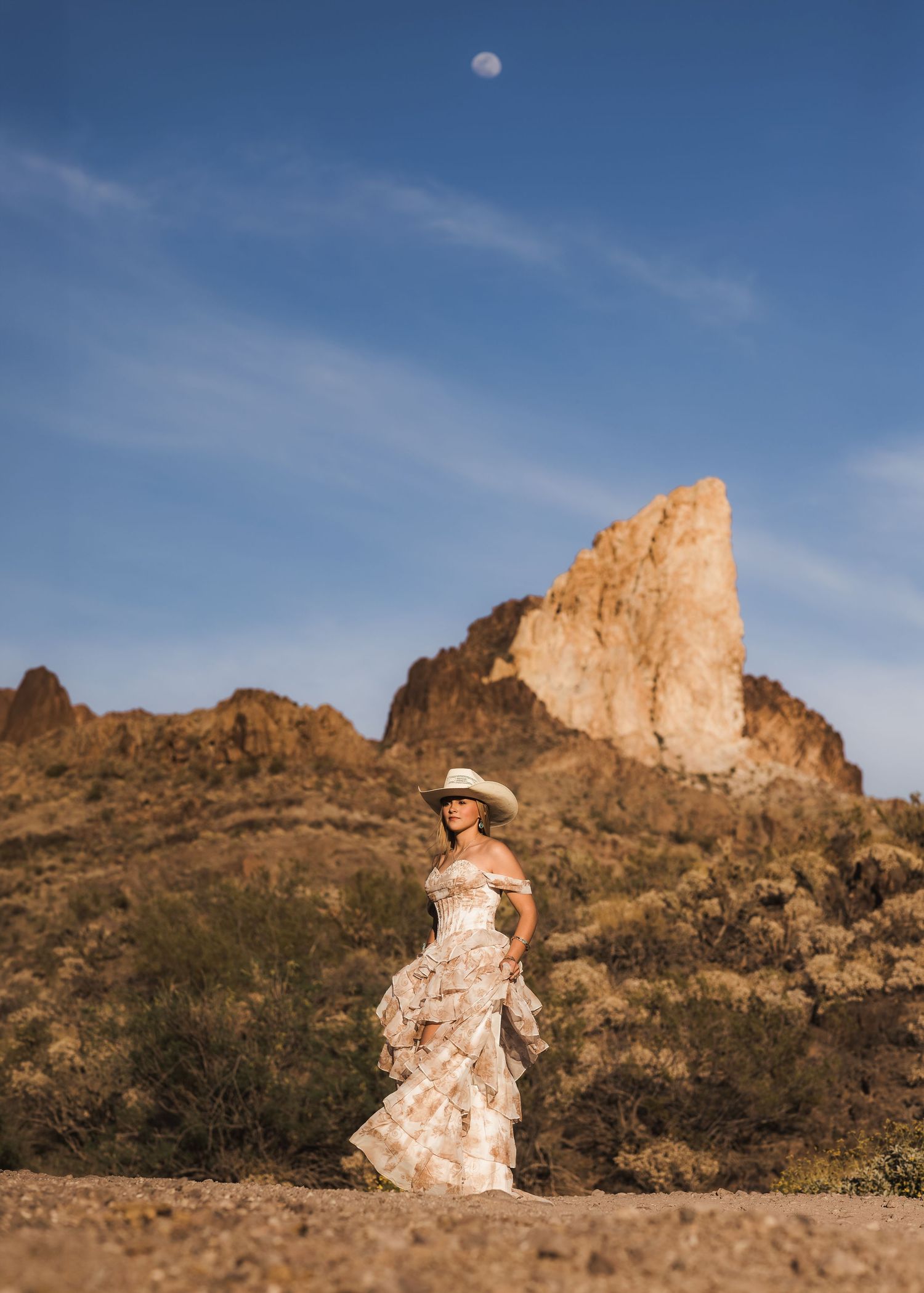 Woman in a ruffled floral dress and cowboy hat standing in a desert landscape under the moon.