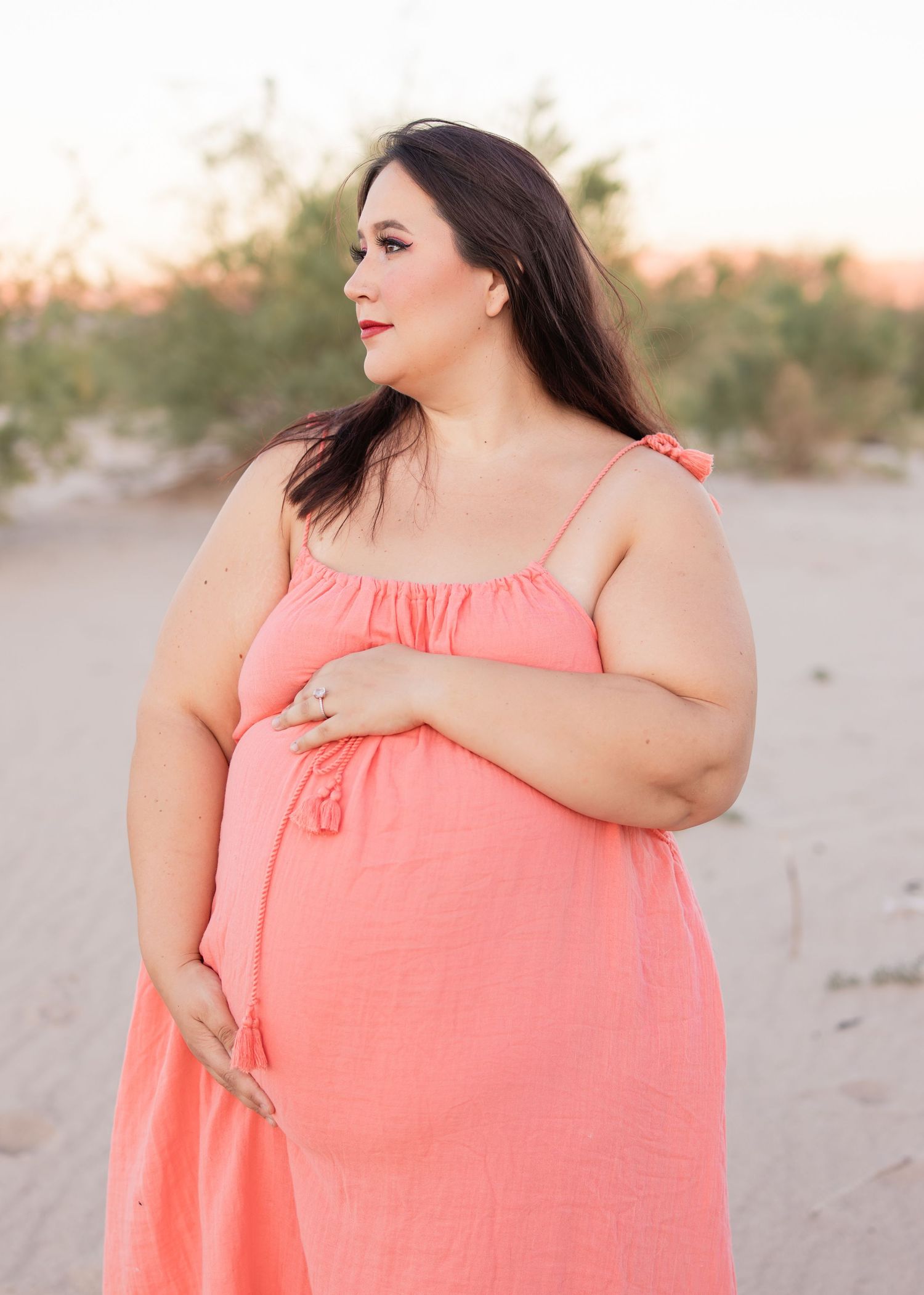 Pregnant woman in coral maxi dress posing outdoors at sunset in a desert landscape.