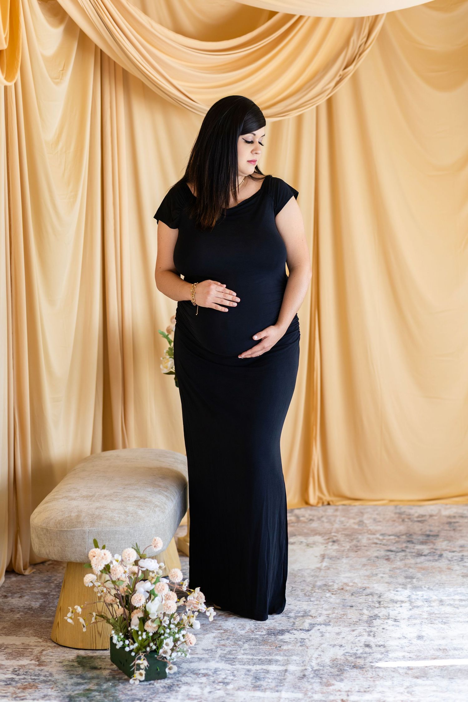 Pregnant woman in elegant black maxi dress poses in studio with gold drape backdrop and flowers.
