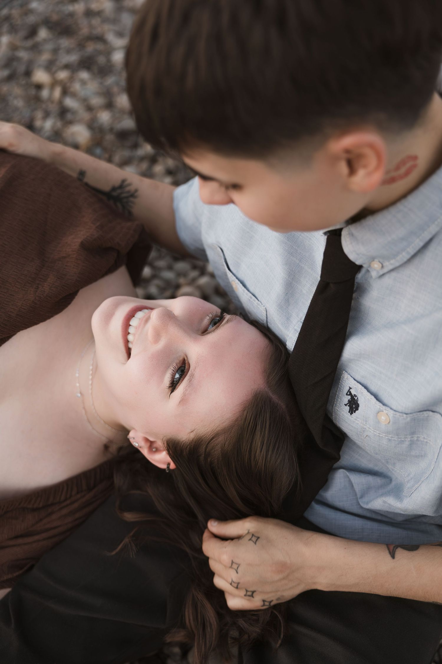 A smiling woman lies on pebbles as a man in a blue shirt leans in close, sharing a tender romantic moment outdoors.