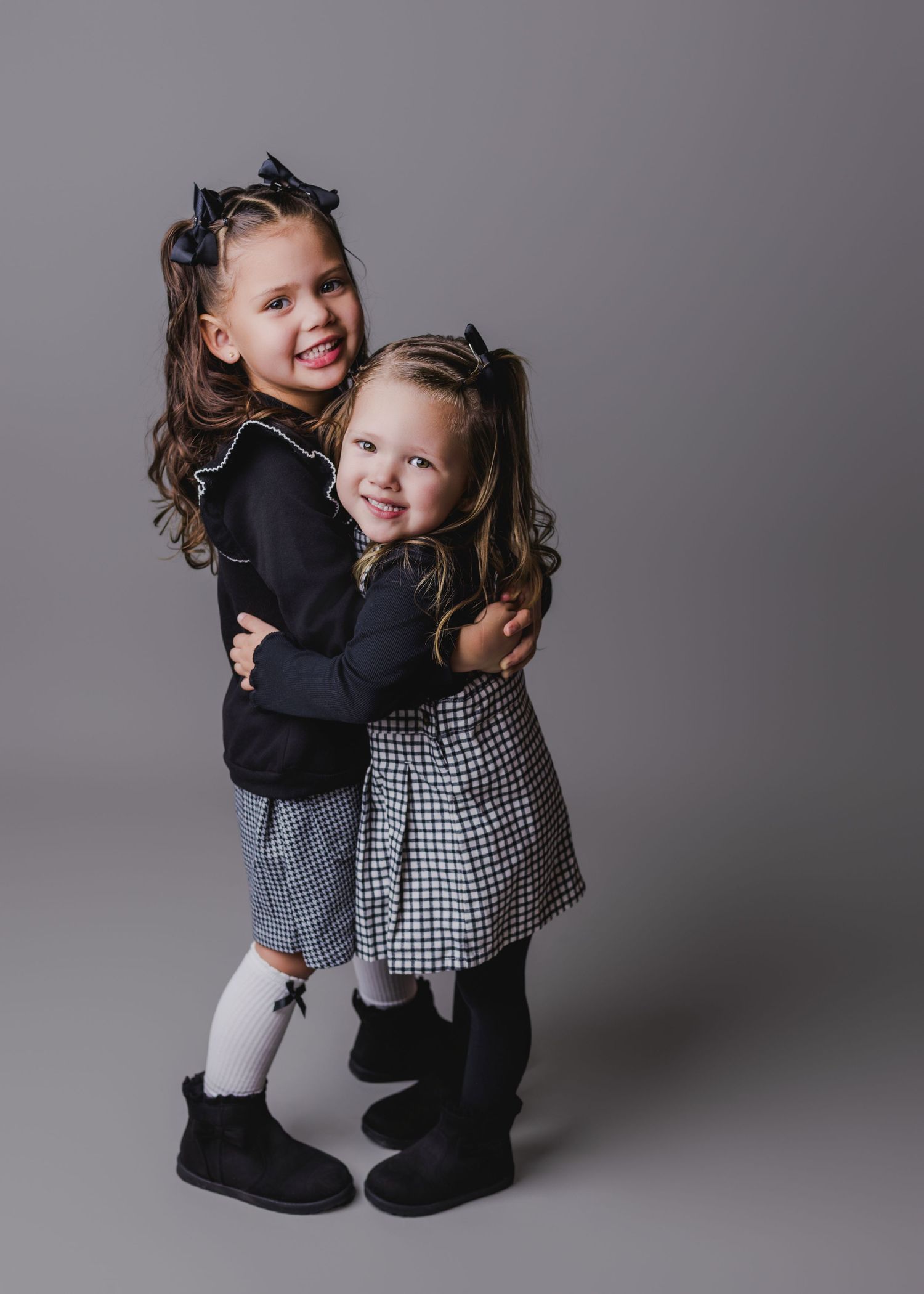 Two young sisters in matching navy outfits with bows hugging and smiling in studio portrait.