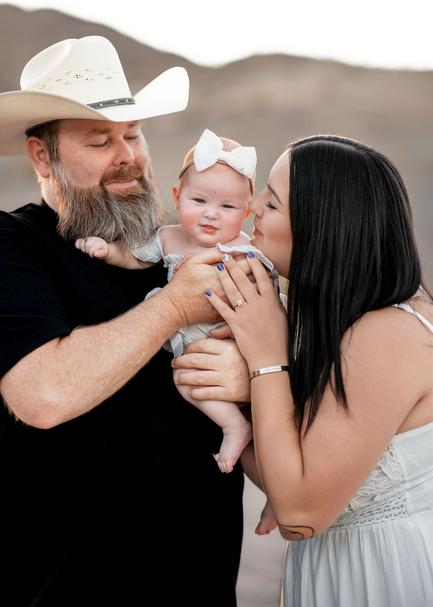 Couple with bearded man in cowboy hat and dark-haired woman kissing their baby girl outdoors at sunset.