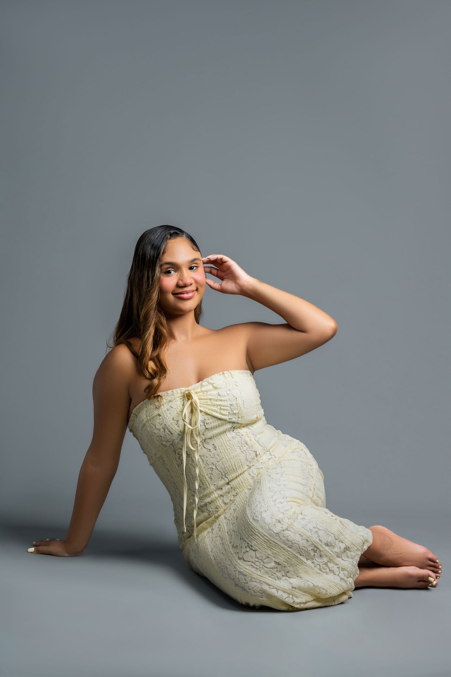A young woman in a cream strapless lace dress sits gracefully on the floor in a studio.