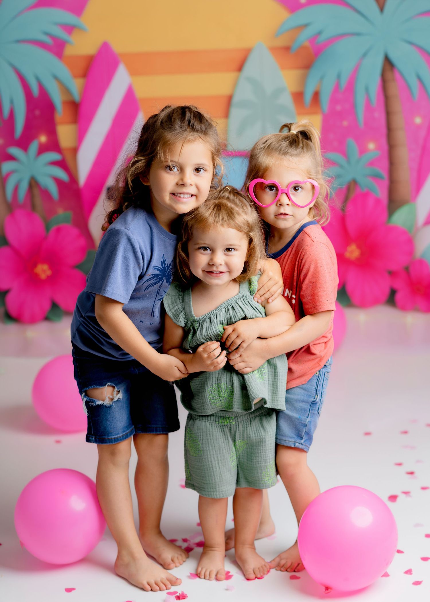 Three young siblings hugging at a colorful tropical-themed birthday photo session.