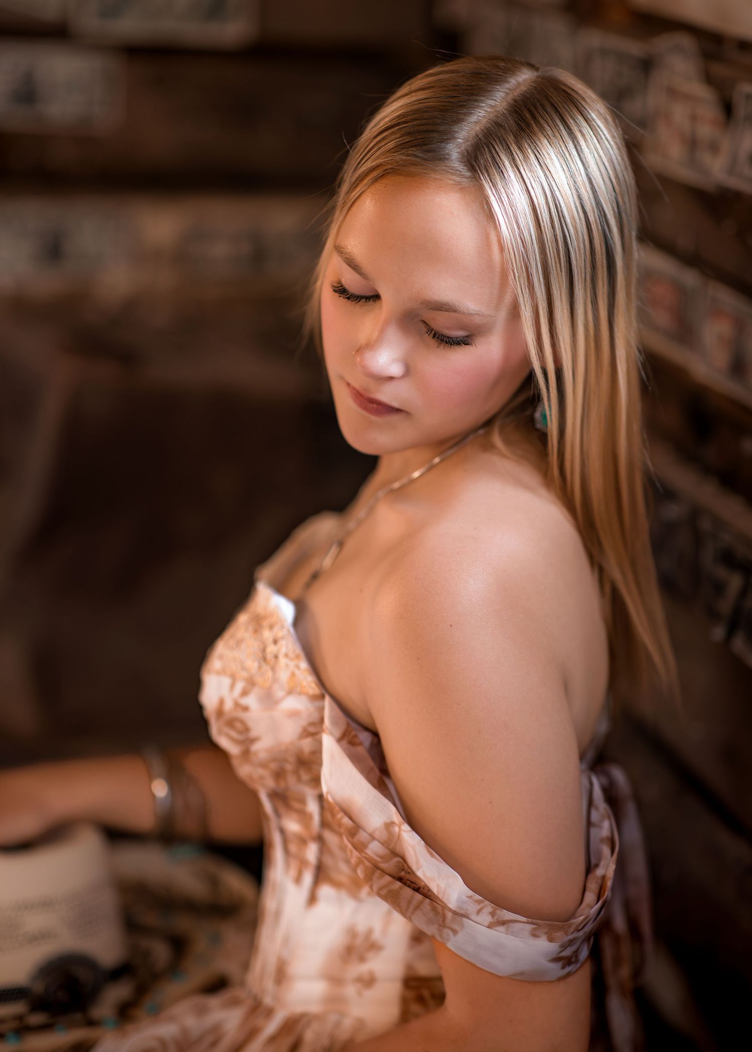Young blonde woman in off-shoulder floral dress poses for elegant senior portrait near rustic brick wall.