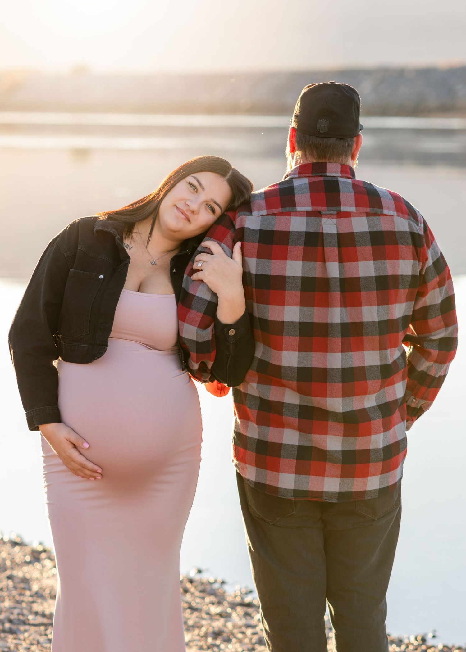 Pregnant woman in pink dress leans on partner in plaid shirt near lake at sunset.