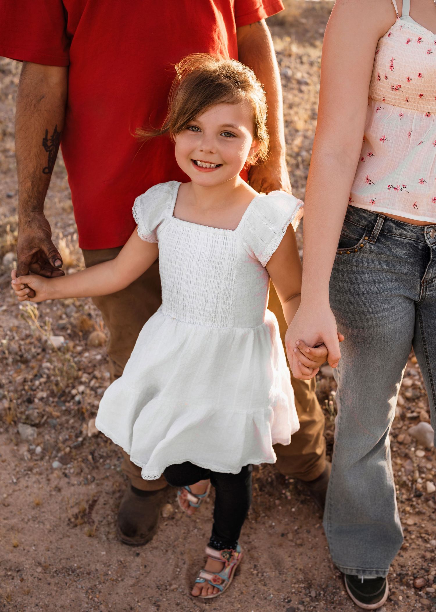 A smiling little girl in a white dress holds hands with family members during an outdoor photo session.