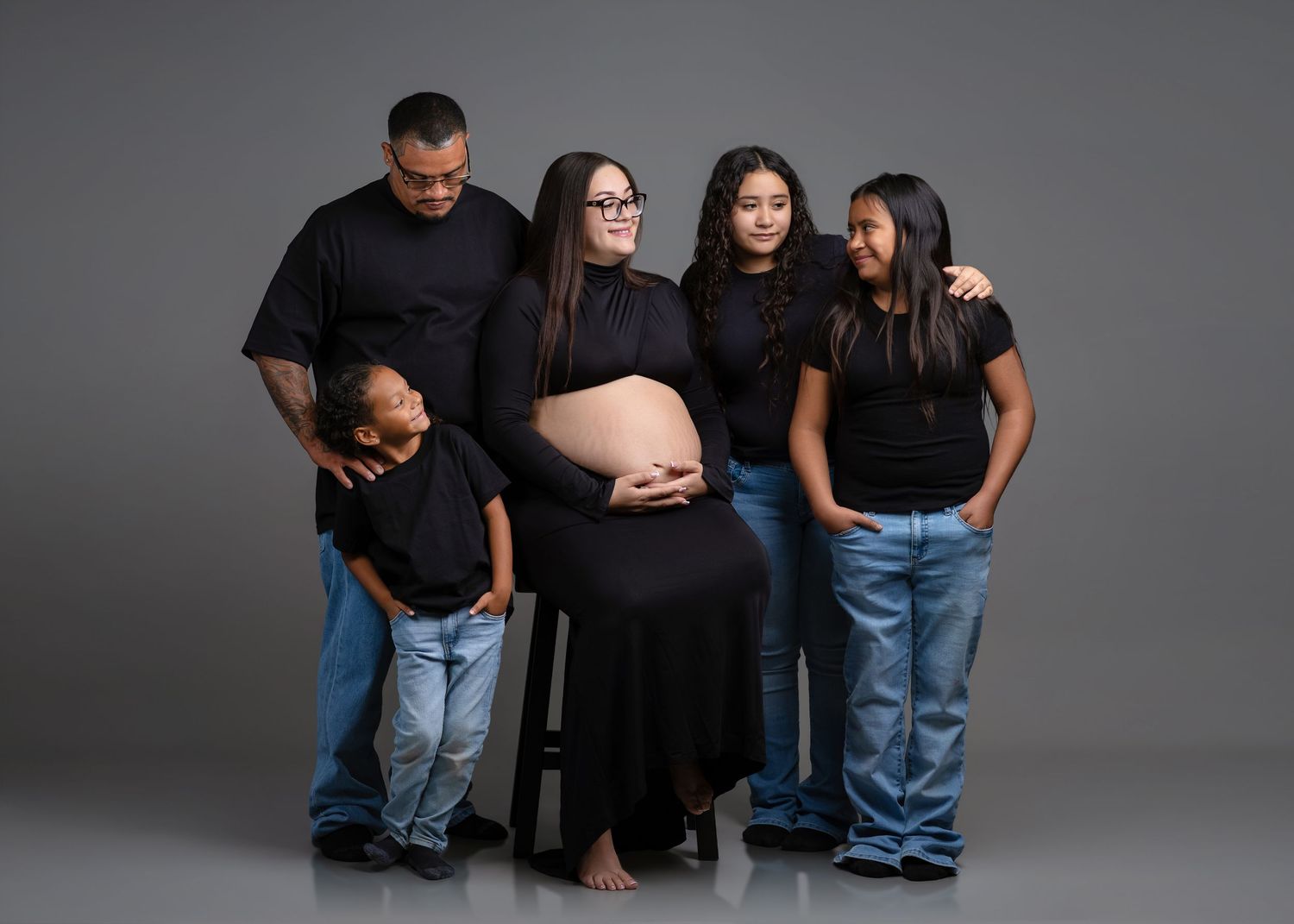 Family of five in black tops and jeans poses together admiring pregnant mothers baby bump in studio.