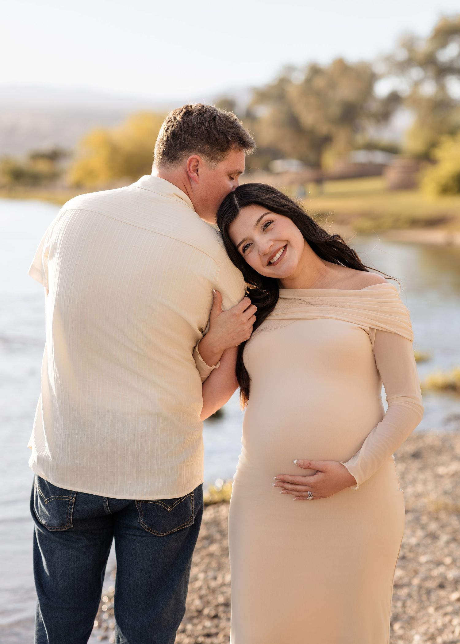 Couple sharing tender moment by riverside, pregnant woman in beige off-shoulder dress smiling warmly.