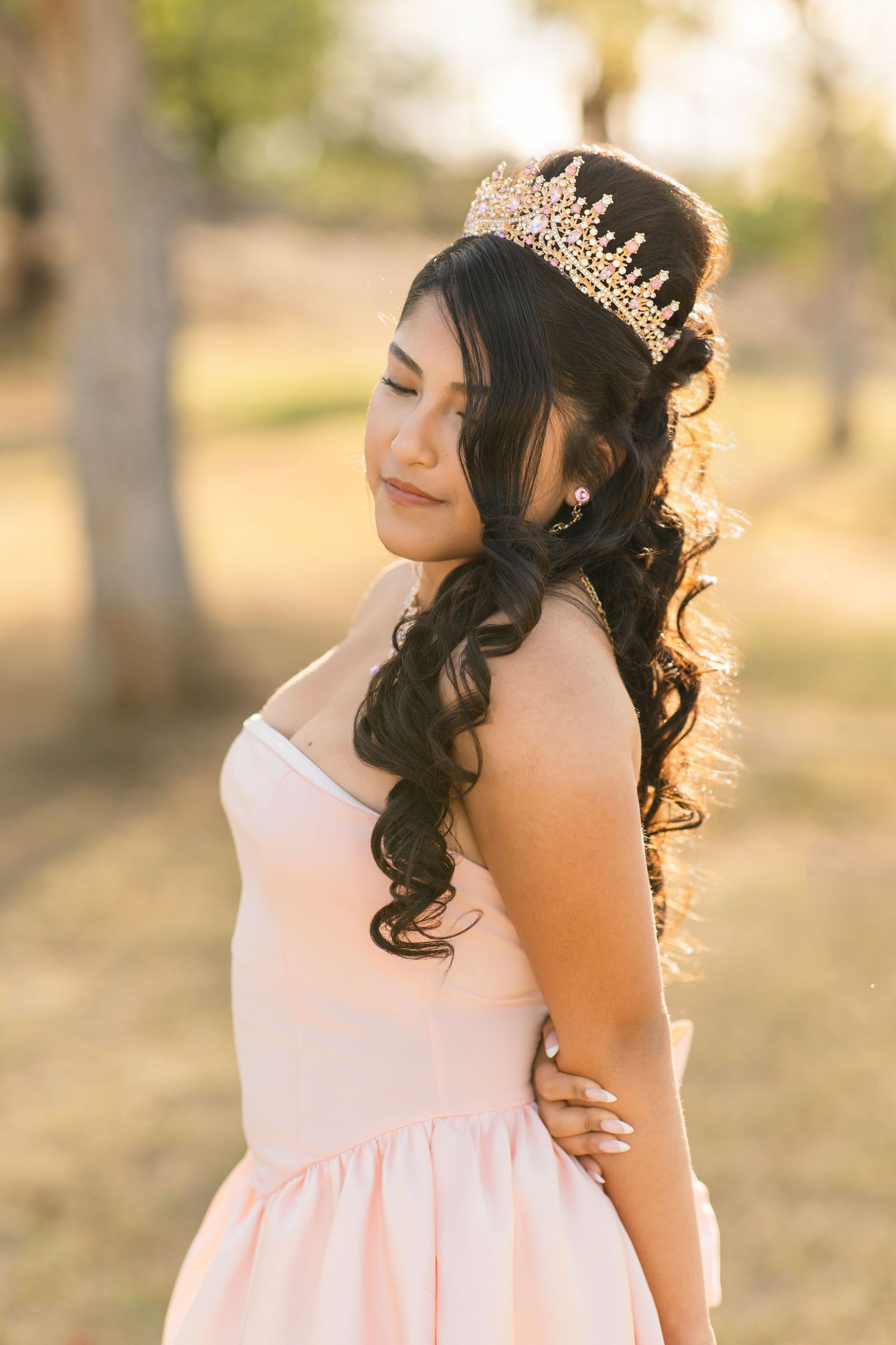 Young woman in pink dress and gold crown tiara poses outdoors during golden hour photography session.