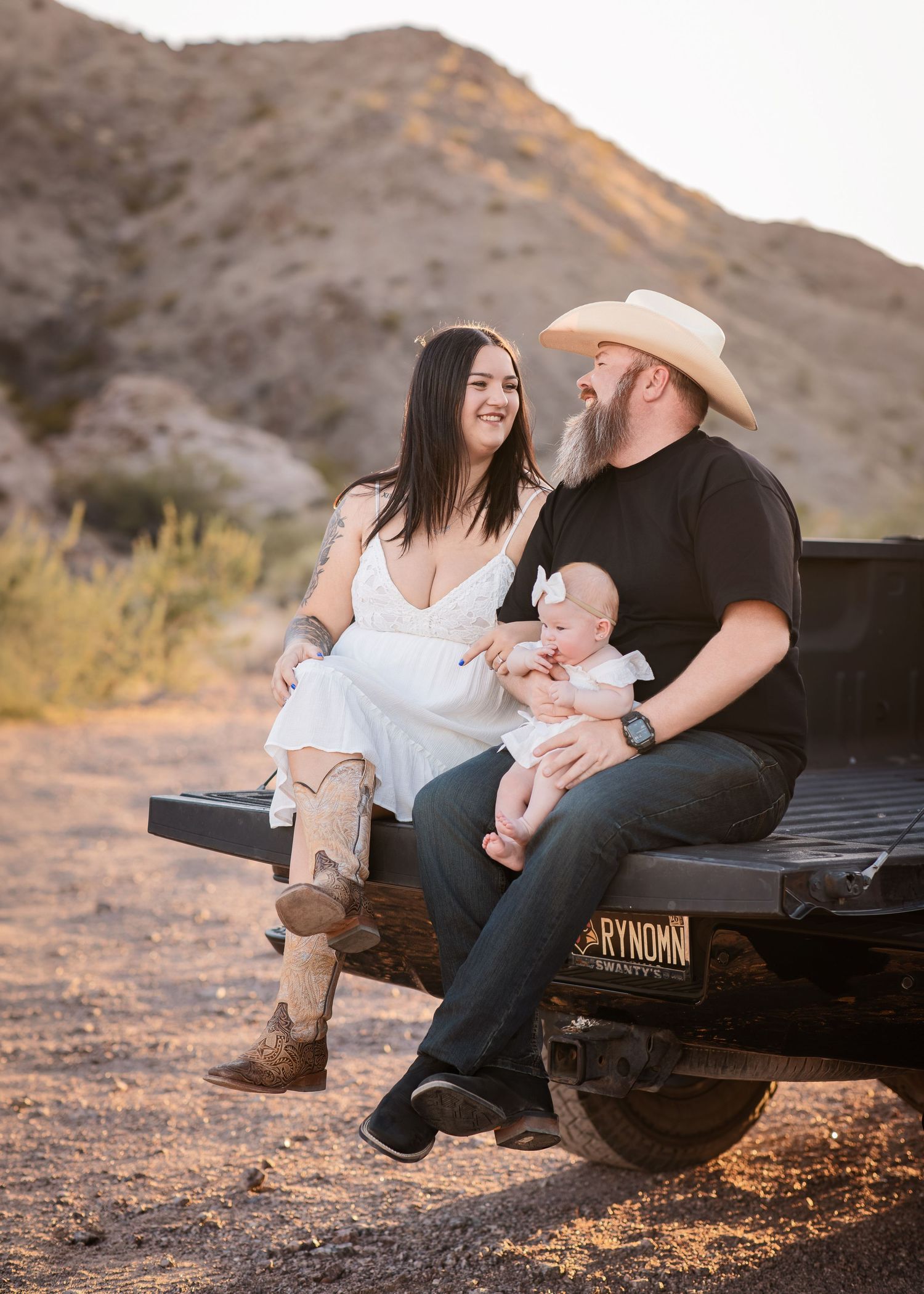 A couple with their baby sits on a truck tailgate in a desert landscape at sunset.