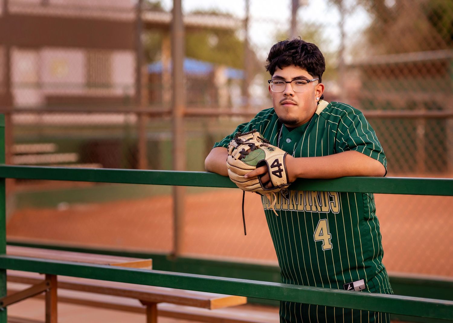 A teenage baseball player in a green uniform leans on a fence holding a glove at an outdoor ballpark.
