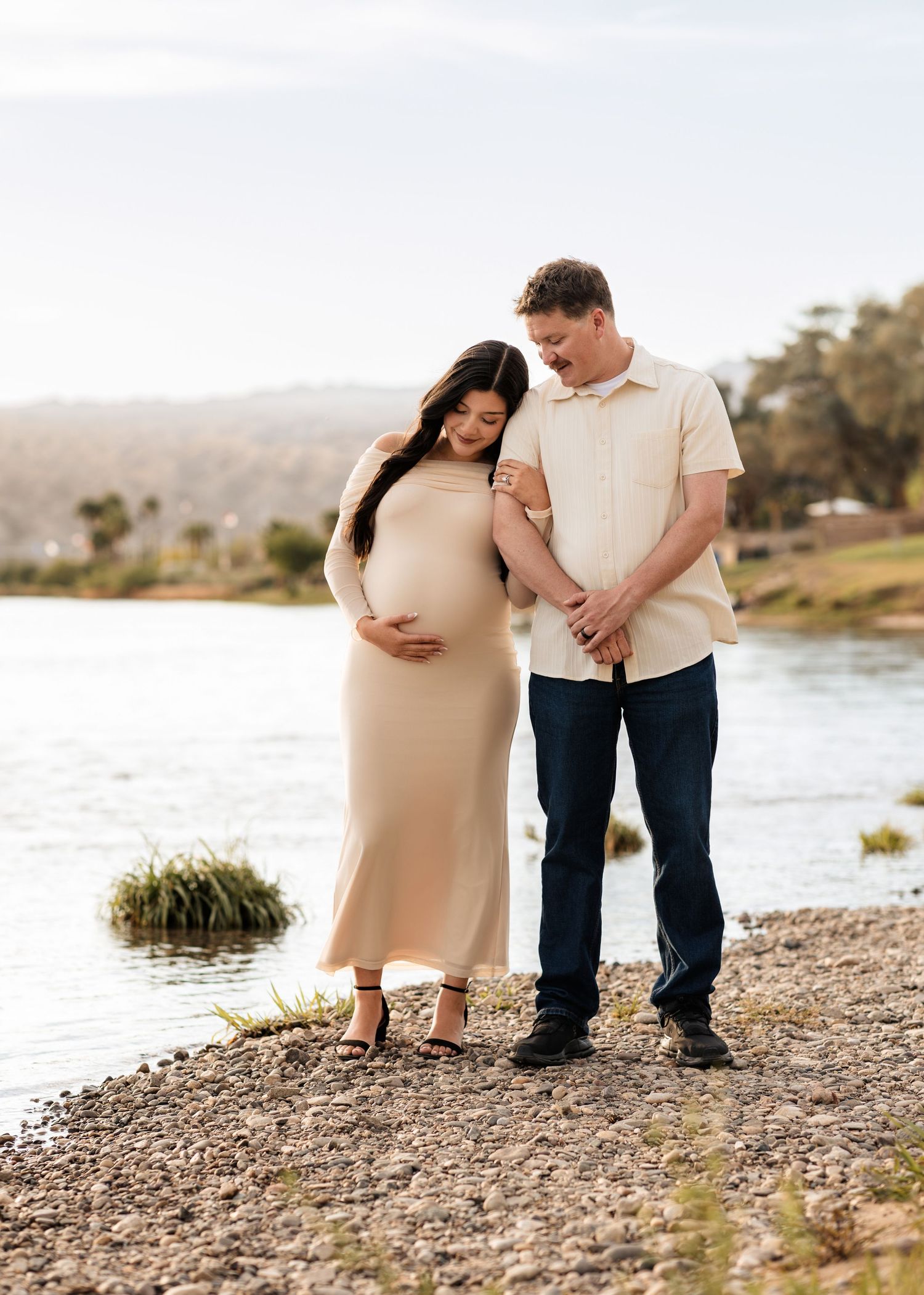Expecting couple shares tender moment by lakeside during outdoor maternity portrait session at golden hour.