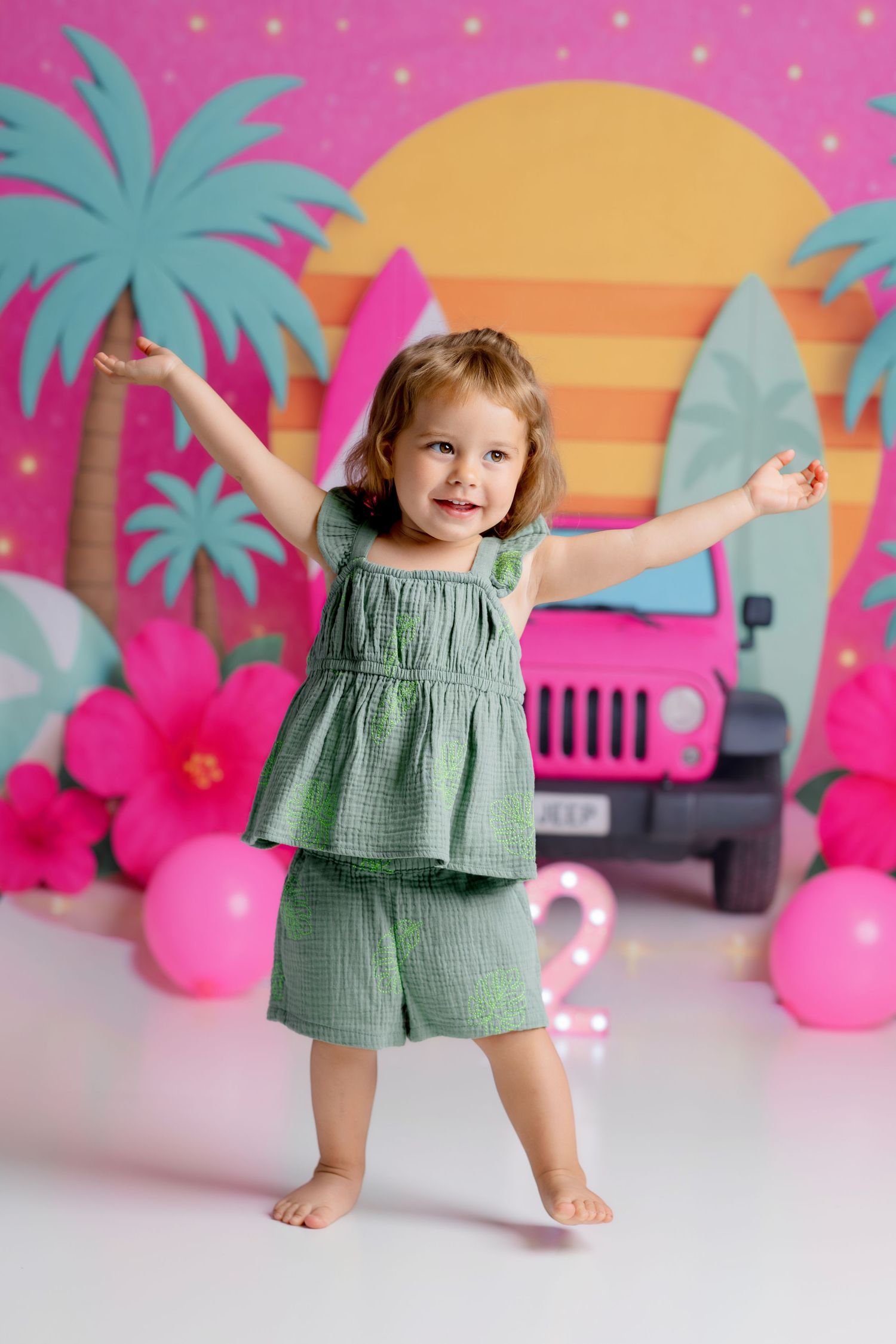 Toddler girl in green outfit celebrates 2nd birthday with tropical Barbie-themed backdrop featuring pink Jeep and palm trees.