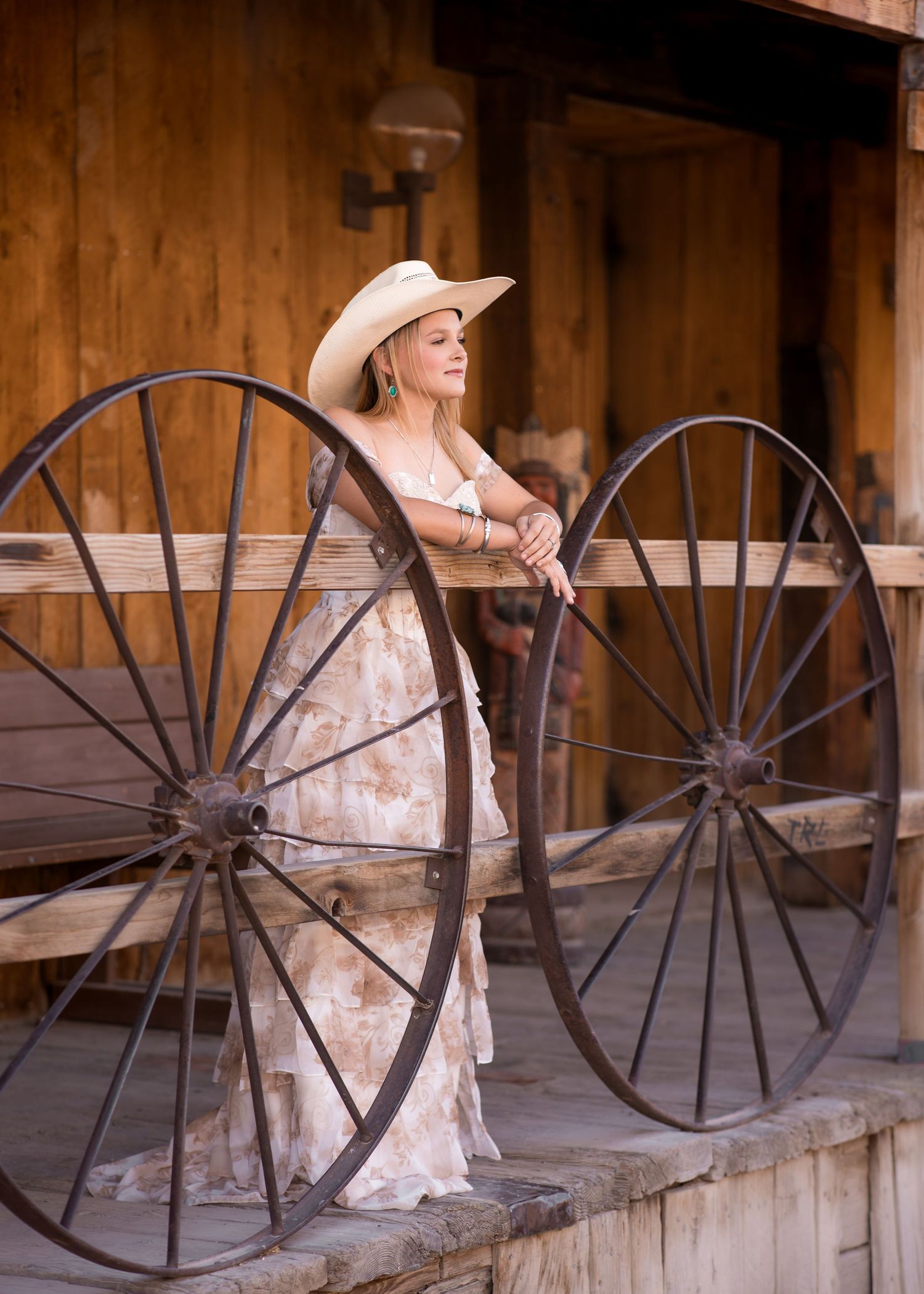 Blonde woman in floral gown and cowboy hat leans on antique wagon wheels at rustic Western venue.