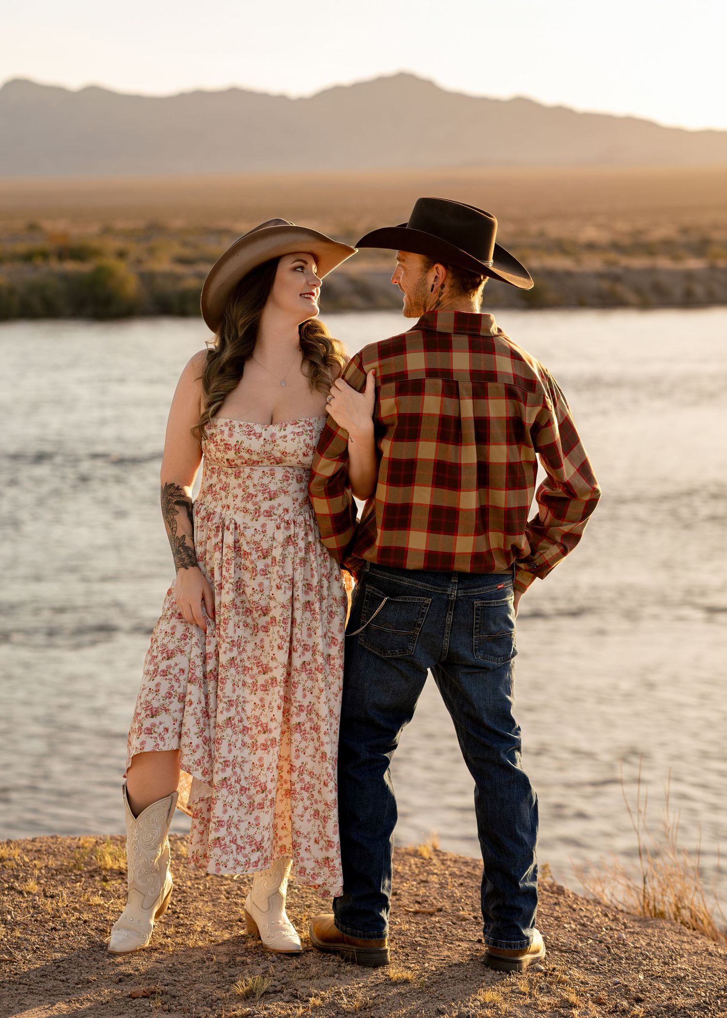 Couple in cowboy hats and western attire standing by a river with mountain views at sunset.
