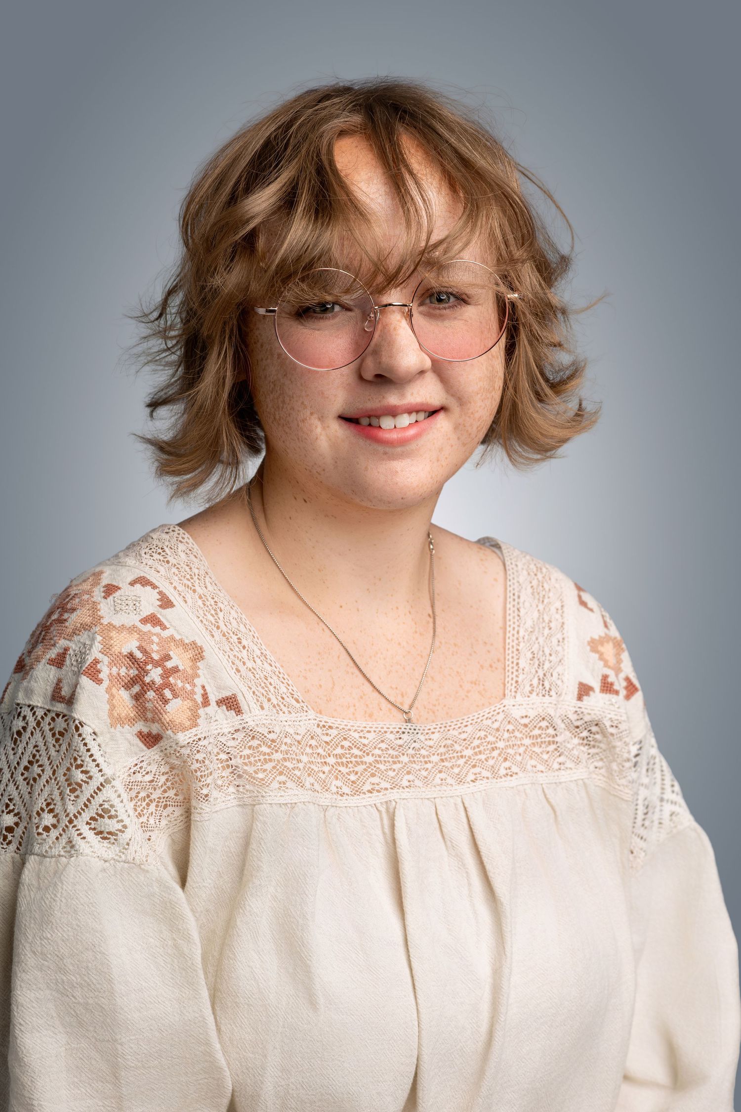 Young woman with short auburn hair and glasses smiles warmly wearing cream embroidered blouse in studio portrait.