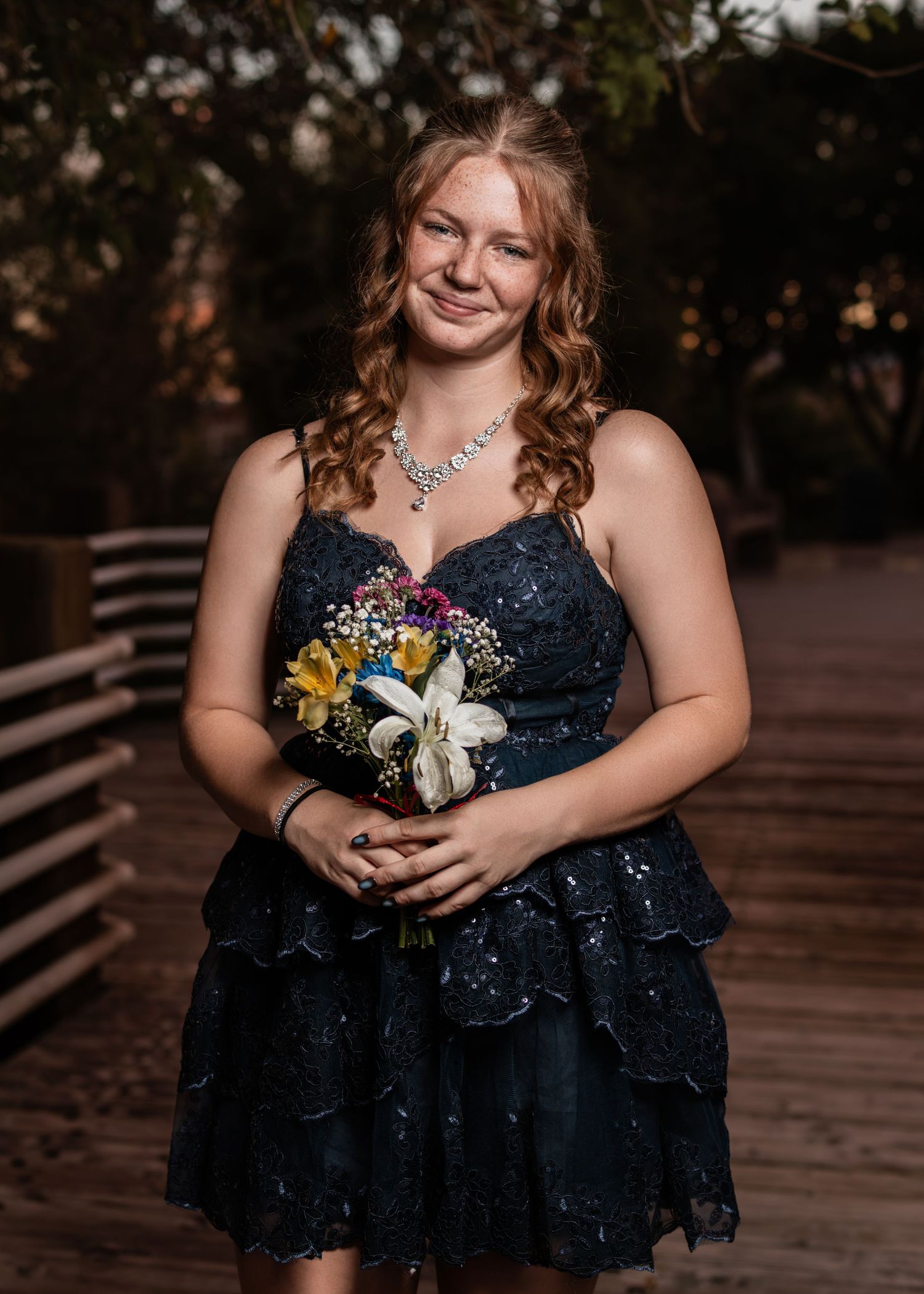 Smiling young woman in navy sequin dress holds colorful bouquet on wooden bridge at dusk.