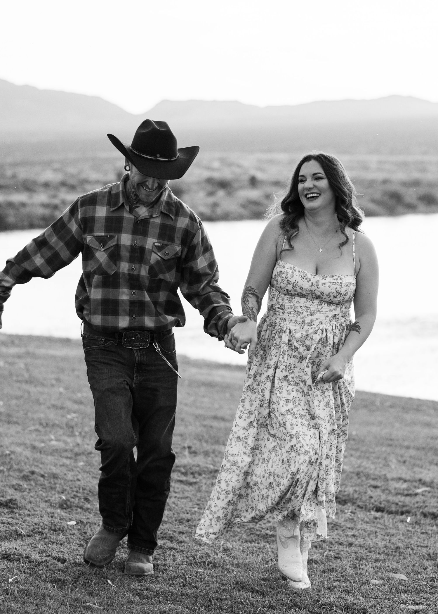 Black and white photo of a couple holding hands and walking near a river in western attire.