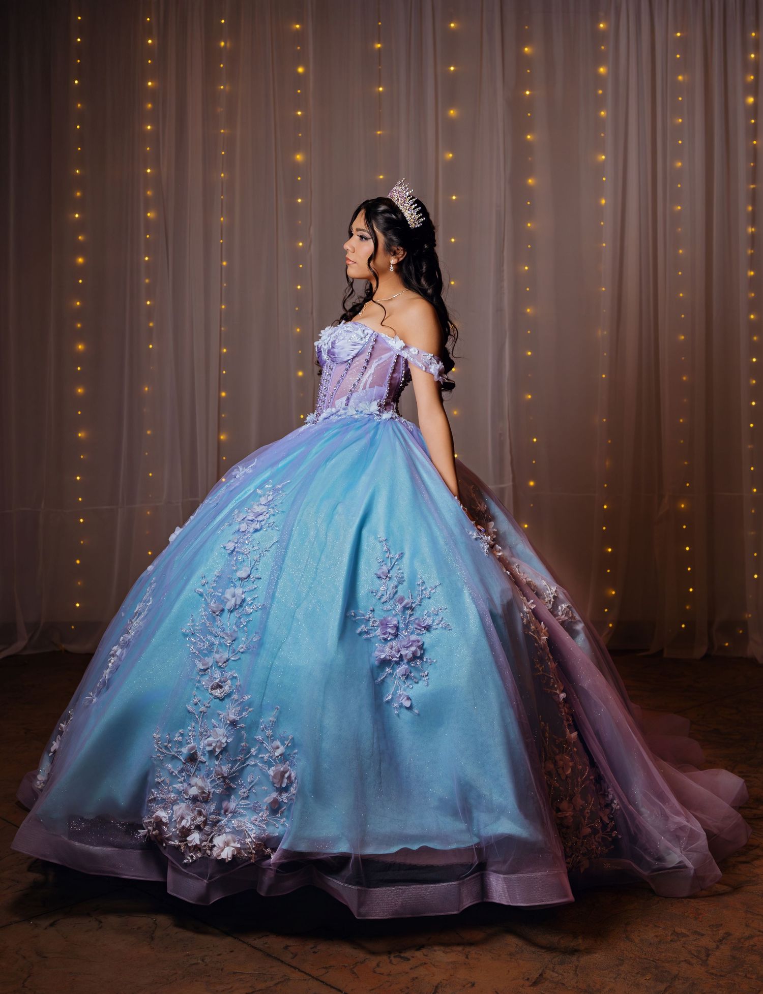Young woman in stunning blue and lavender quinceañera ball gown with floral details and tiara.