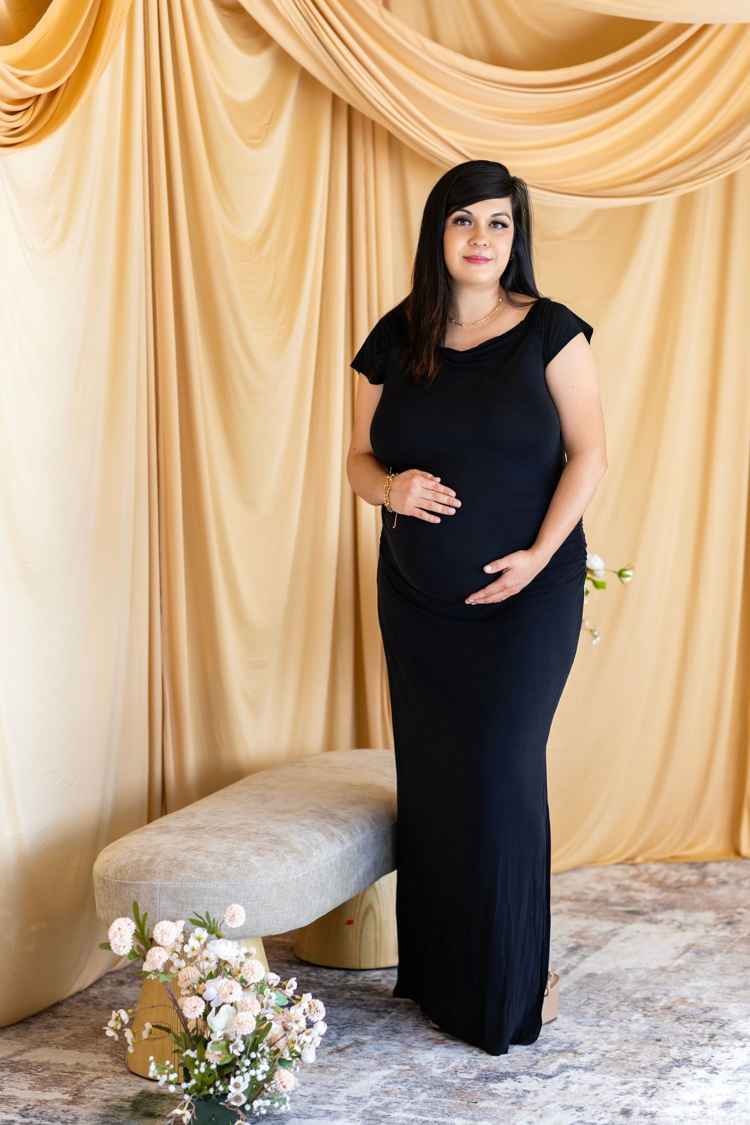 Pregnant woman in black maxi dress smiles warmly in studio maternity photo with gold curtain backdrop.