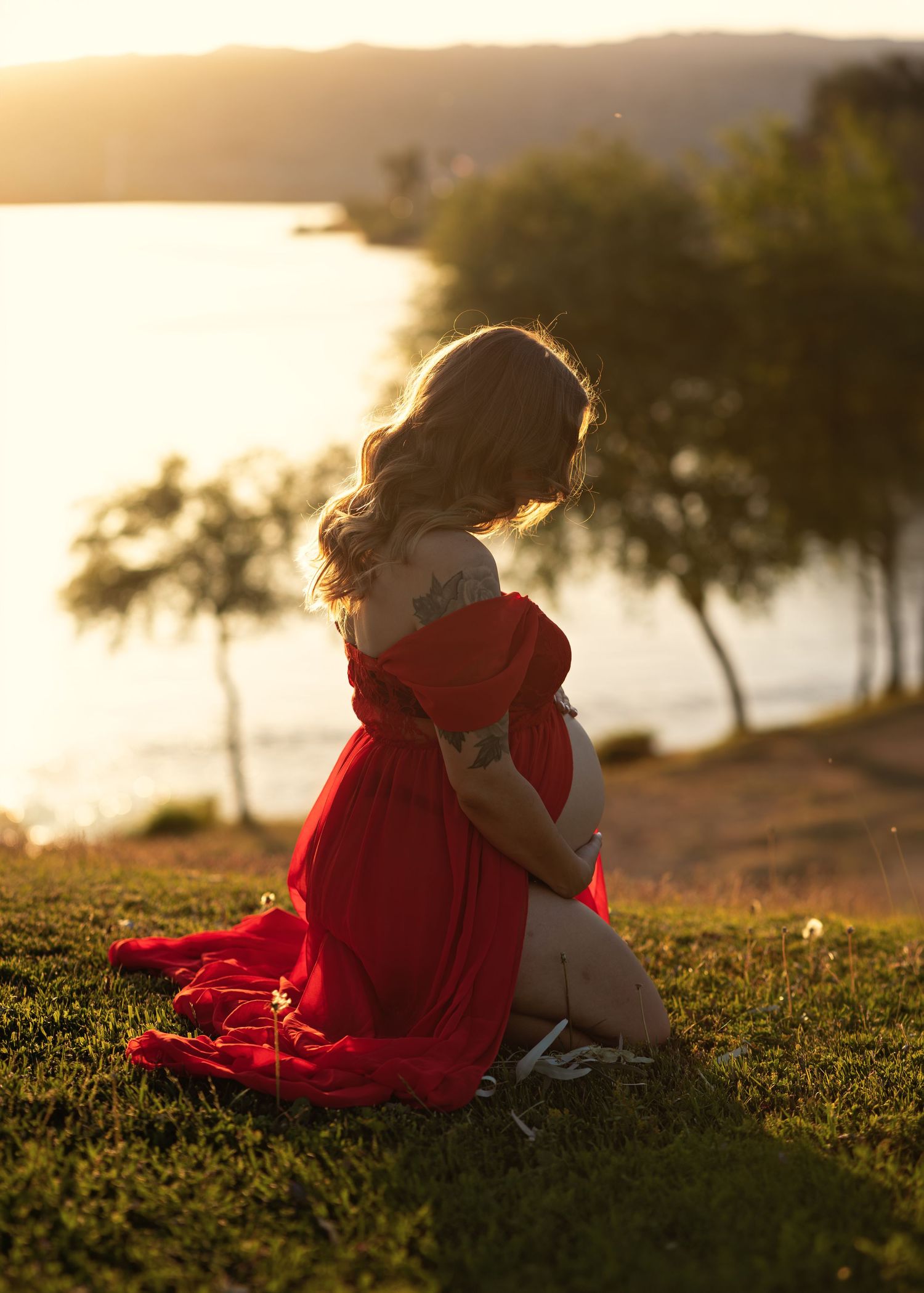 A pregnant woman in a red off-shoulder gown sits on grass at golden hour by a lake.