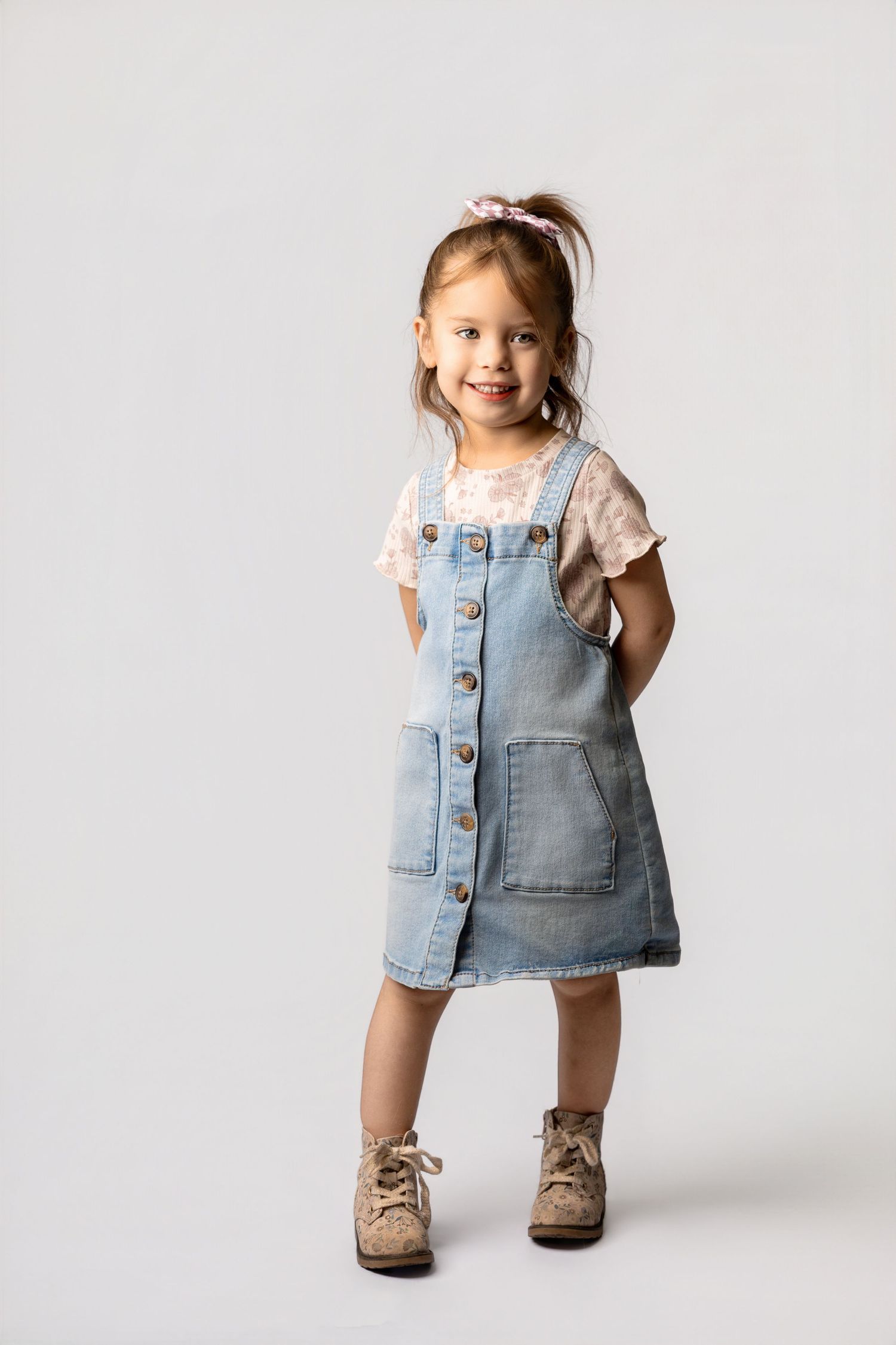 Smiling little girl wearing a denim pinafore dress, floral shirt, and boots, posing against a white background.