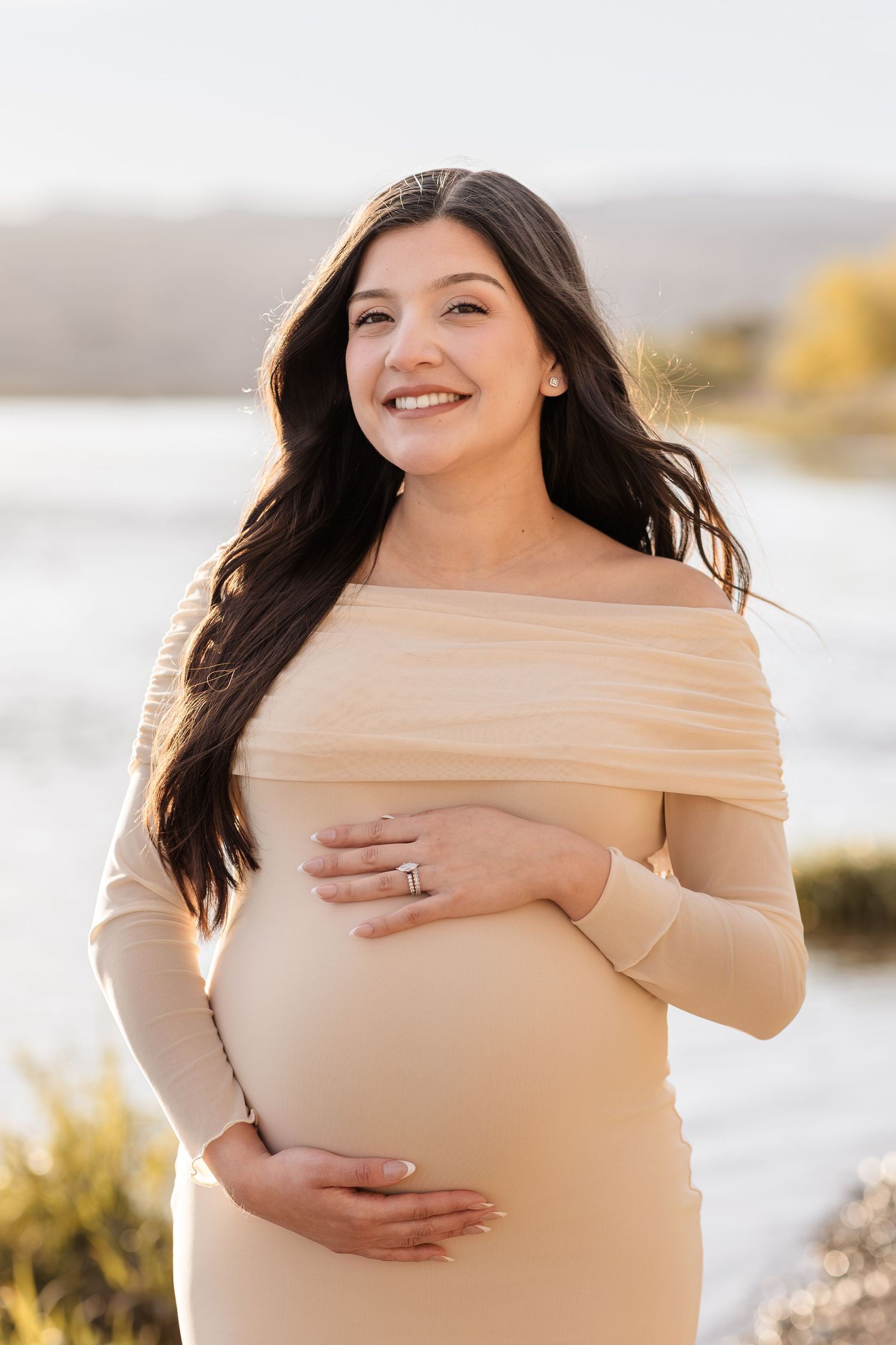 Smiling pregnant woman in off-shoulder beige dress cradling baby bump outdoors in golden sunlight.