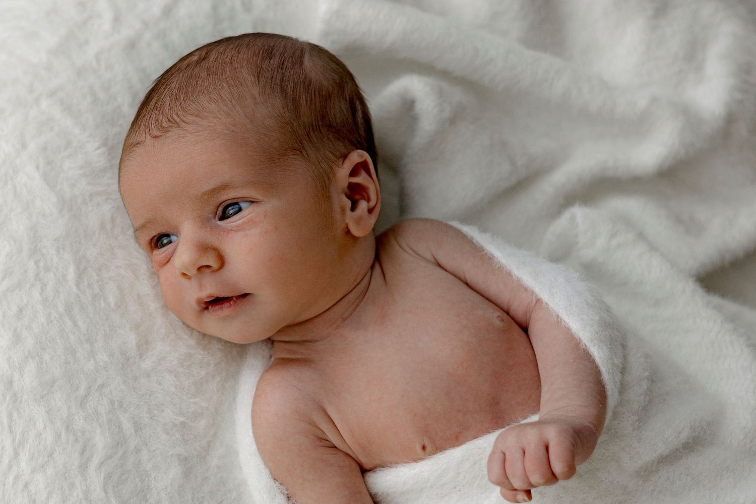 Newborn baby lying on a soft white blanket, looking alert with big eyes in a peaceful nursery setting.