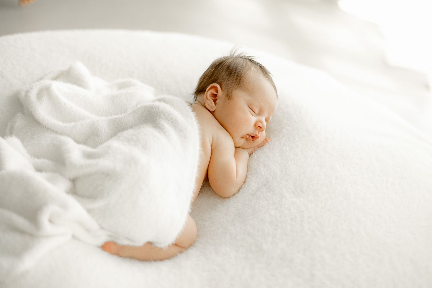 Peaceful newborn baby sleeping on white fluffy surface, wrapped in soft white blanket, serene infant portrait.
