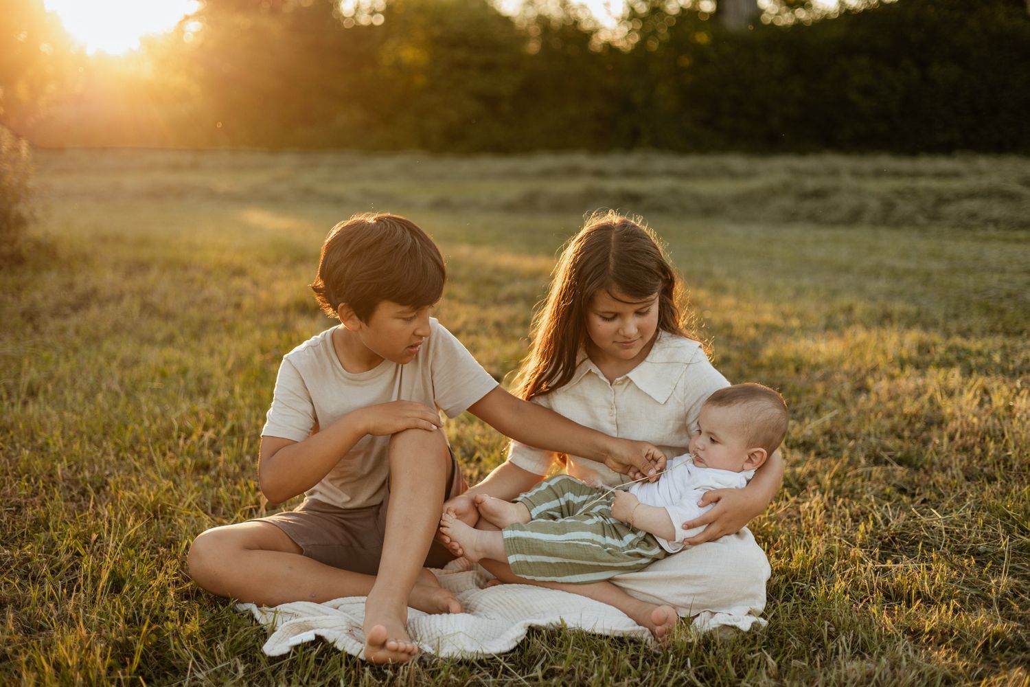 Two children and a baby sitting on a blanket in a sunny green field during golden hour sunset.