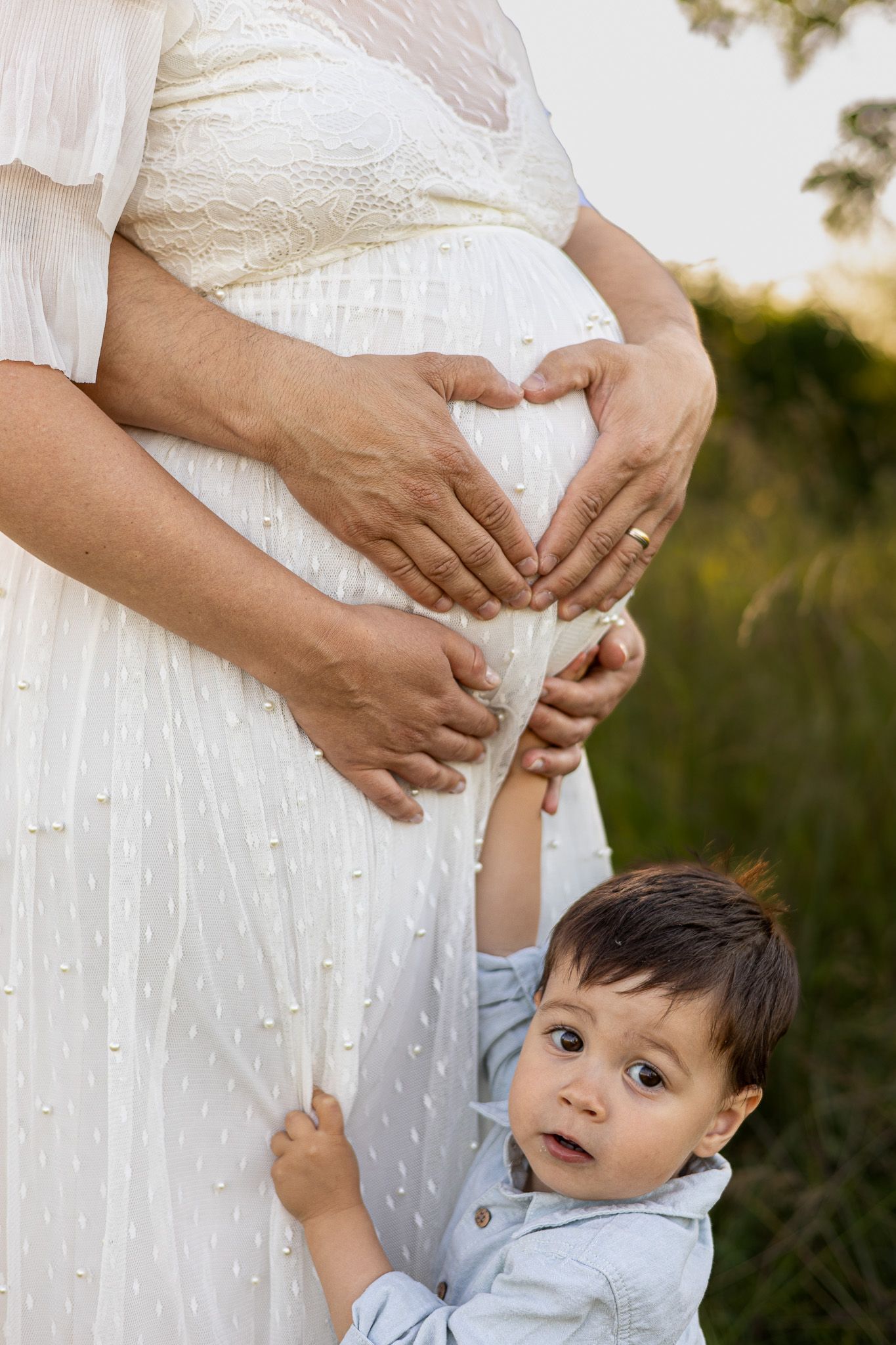 Pregnant woman in white dress with toddler boy making heart shape on baby bump outdoors.