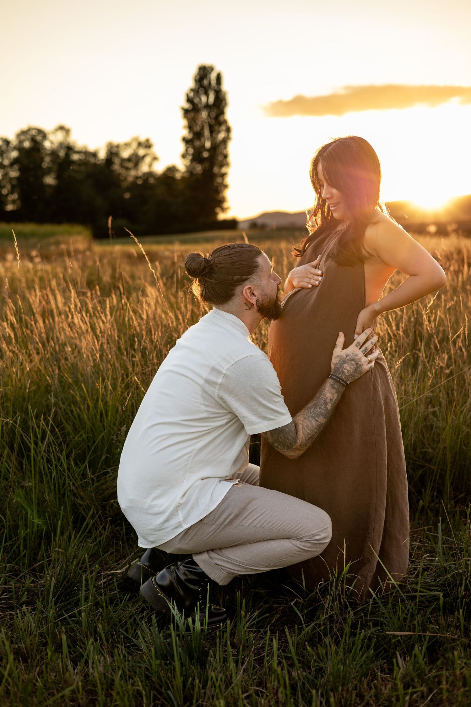 Man kneeling in golden field at sunset, tenderly touching pregnant woman's belly during maternity photo session.