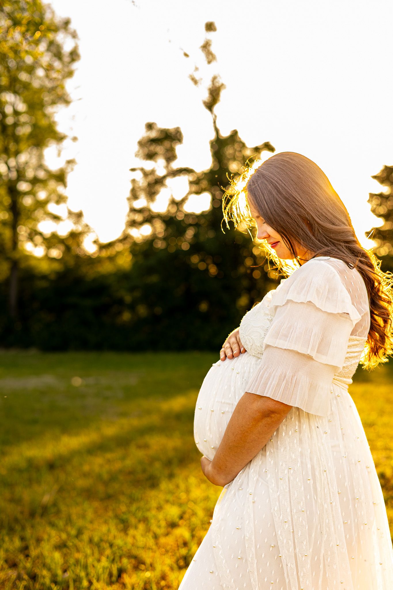 Pregnant woman in white dress cradling baby bump outdoors at golden hour sunset in lush green park.