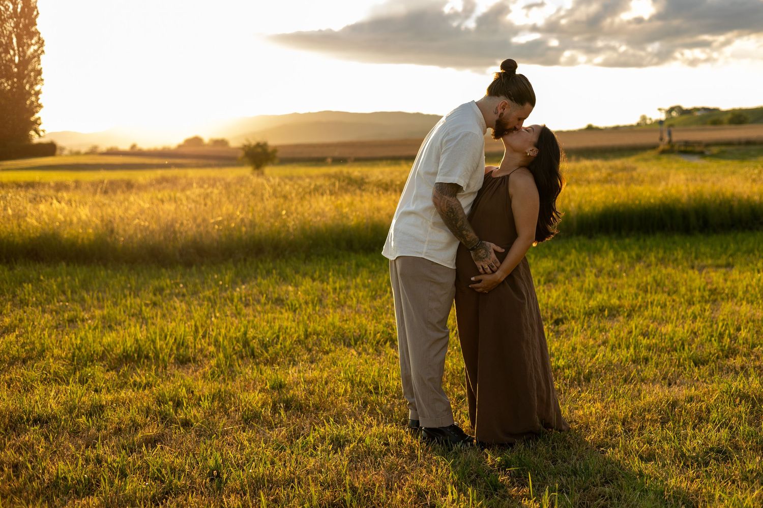 Couple sharing a tender kiss in a golden sunlit field at sunset, with mountains and open farmland in the background.
