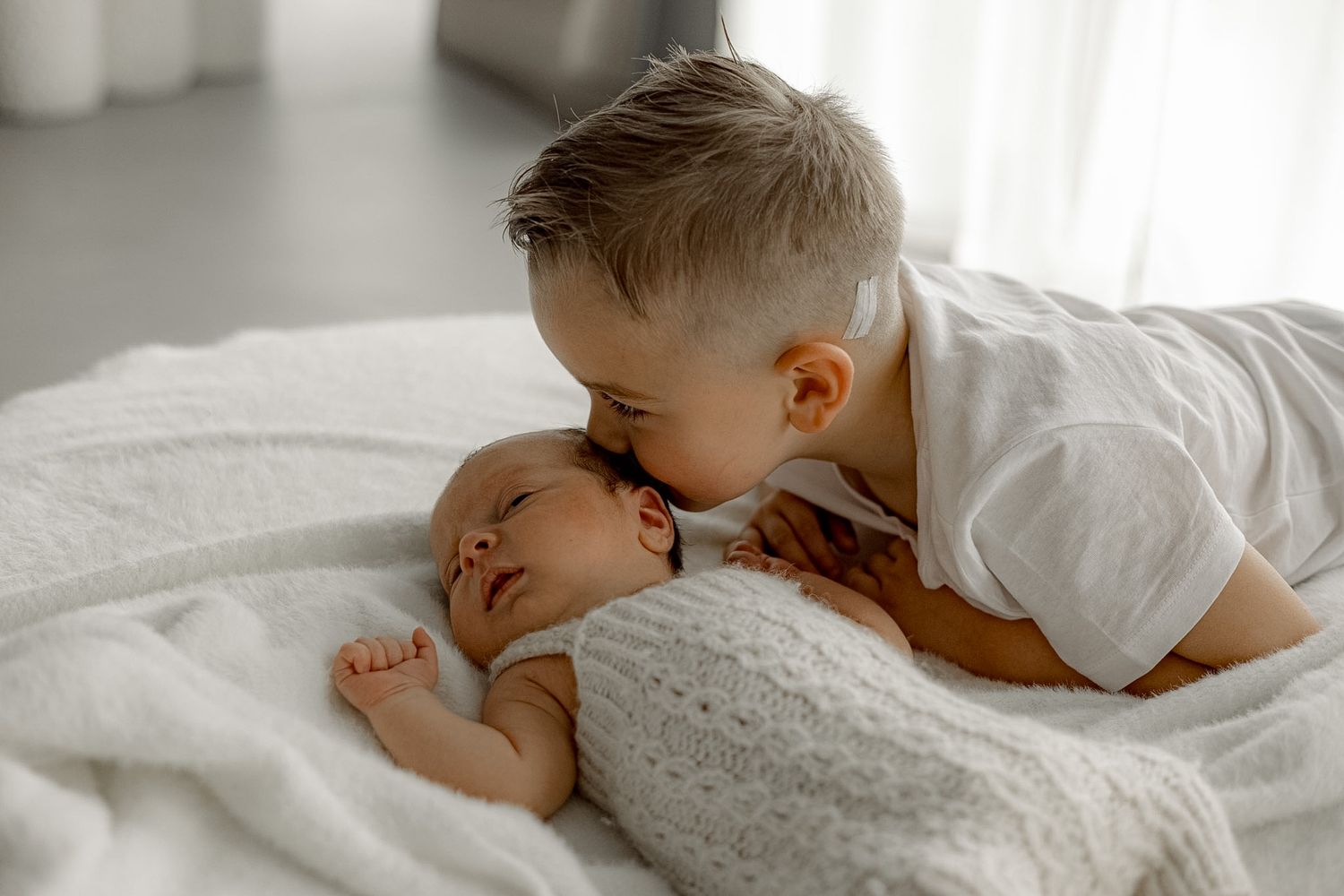 A young boy gently kisses a sleeping newborn baby wrapped in a white blanket on a soft white bed.