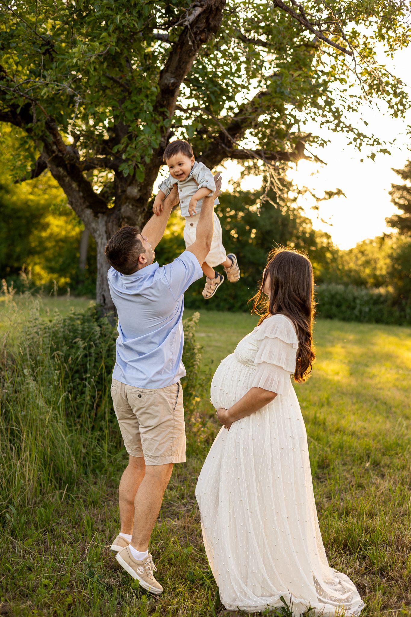 A happy family outdoors at golden hour, father lifting toddler while pregnant mother in white dress smiles nearby.