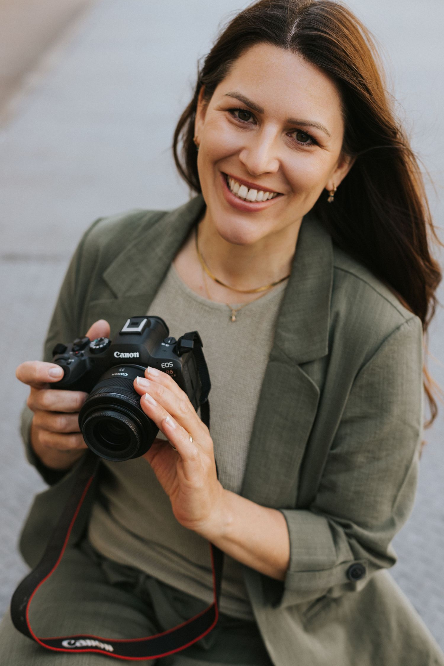 Smiling woman in olive blazer holding a Canon EOS camera outdoors, ideal for photography or lifestyle content.