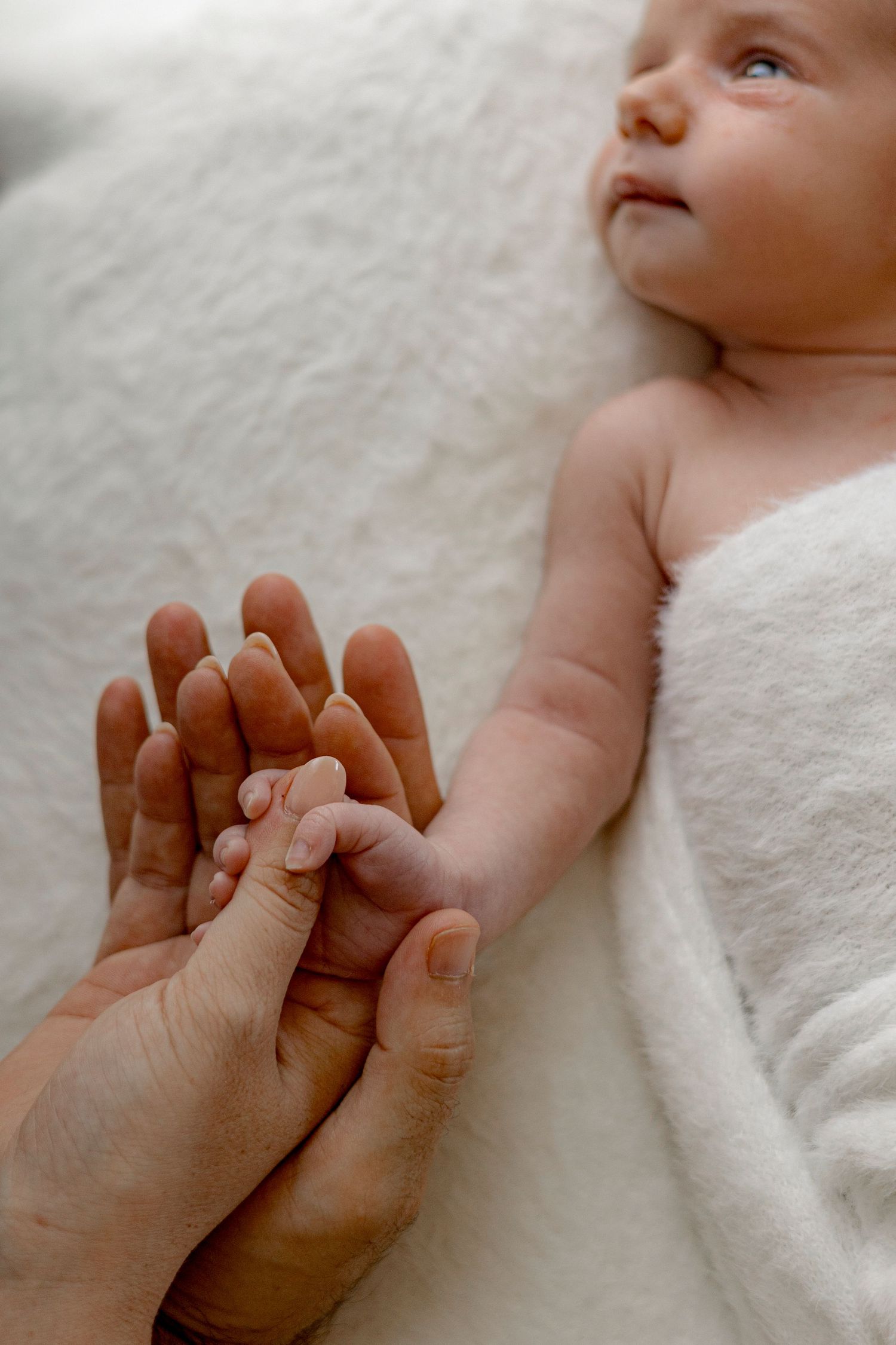 Newborn baby lying on white blanket gently holding adult fingers, showcasing the tender bond between parent and child.